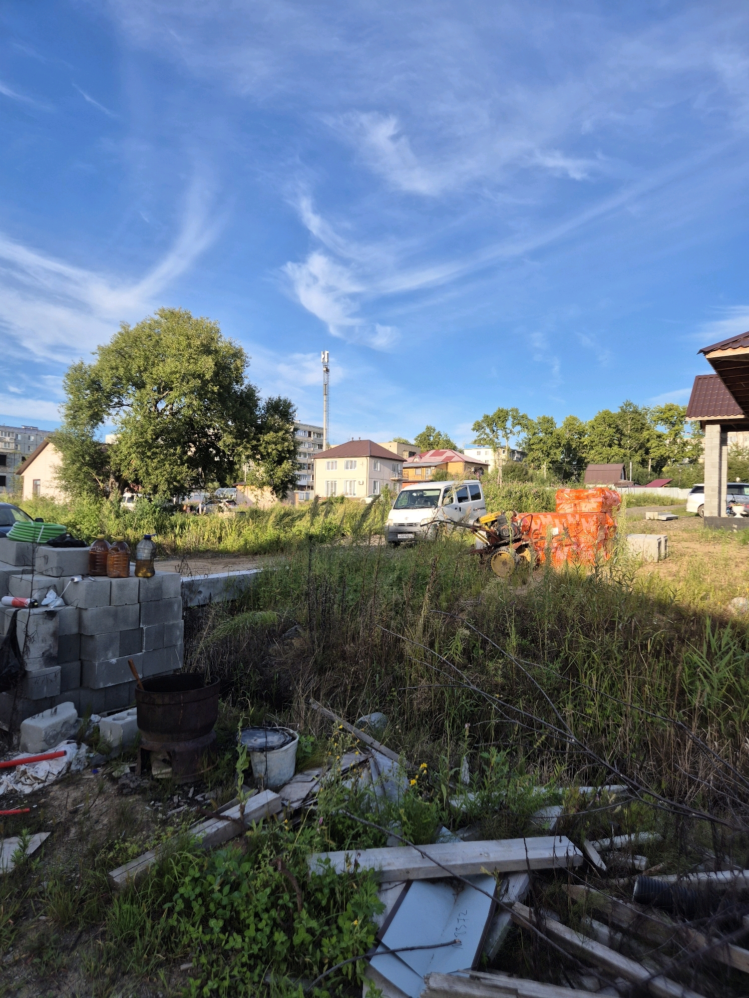 A low-angle outdoor daytime shot features a construction site in a somewhat overgrown area. In the foreground, to the left, is a stack of gray concrete blocks. To the right of the blocks, an old, dark metal pot sits on a makeshift stand, with a white bucket to its right. Beyond this, the ground is covered in tall, dry grass and various debris, including wooden planks and what appears to be corrugated plastic. In the middle ground, a white van is parked, and next to it, a small yellow agricultural machine with an orange tarp covering is visible. Further back, a large, leafy green tree stands prominently, with several residential buildings and a cell tower visible behind it. The sky is a bright blue with wispy white clouds.