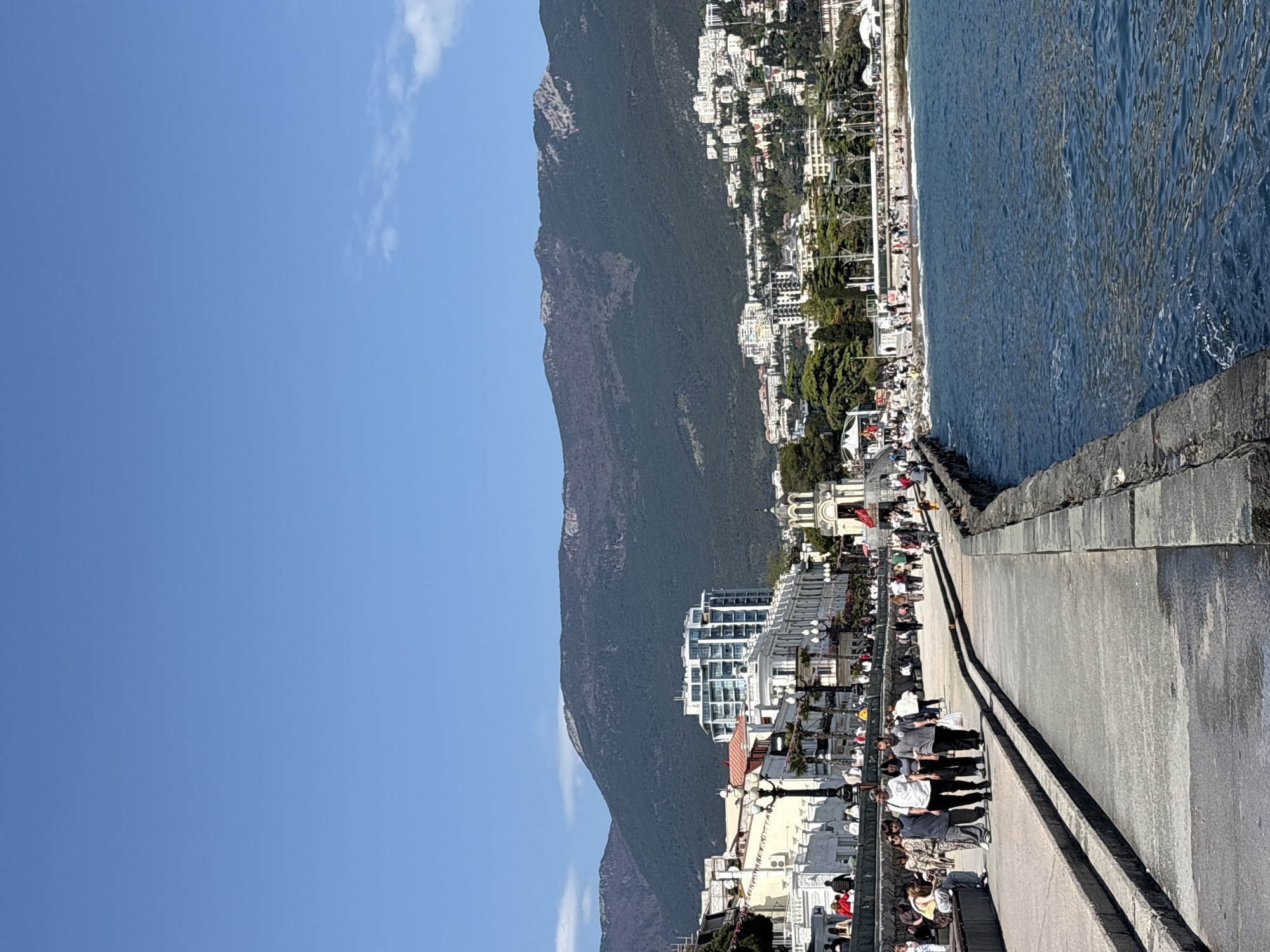 The image depicts a pier in the city of Yalta, Ukraine. A mountain rises in the background, with buildings lining its lower slopes. Along the pier, people stroll and gather. The sea is visible on one side of the pier, with its surface reflecting the sunlight. The sky is clear and blue, with a few scattered clouds. The pier itself is constructed of concrete, with a barrier separating pedestrians from the sea. Overall, the scene presents a view of a bustling pier on a bright day.