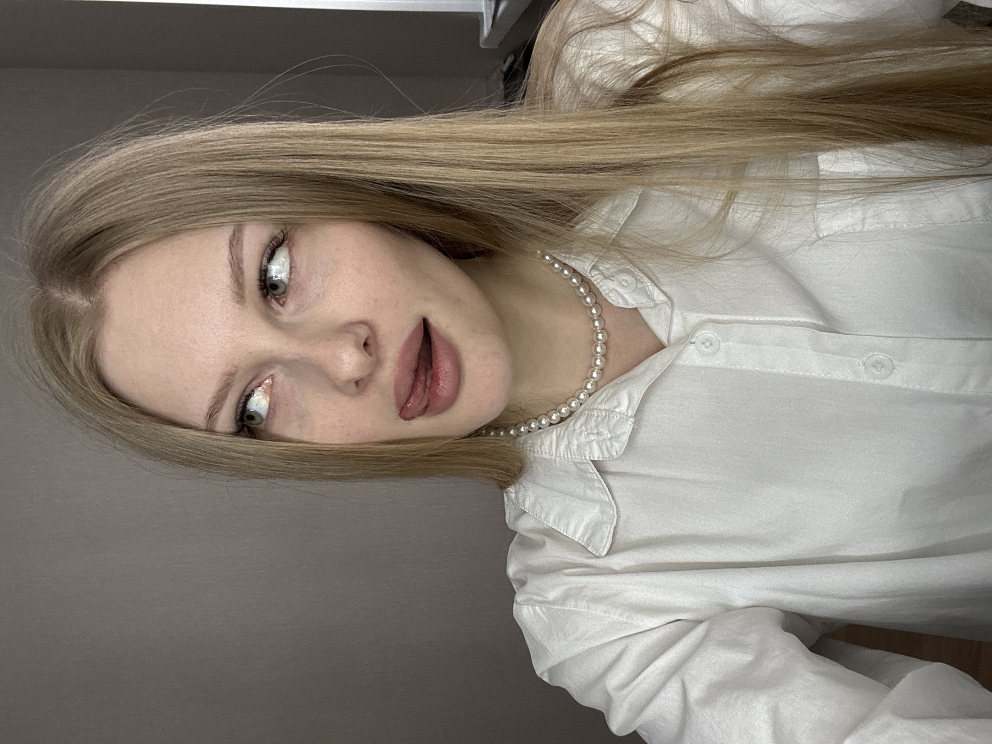 A young woman with long blonde hair, wearing a white button-down shirt and a pearl necklace, is sticking out her tongue. The location is inferred from metadata, indicating the city of Perm, Russia. The background is a plain gray wall.