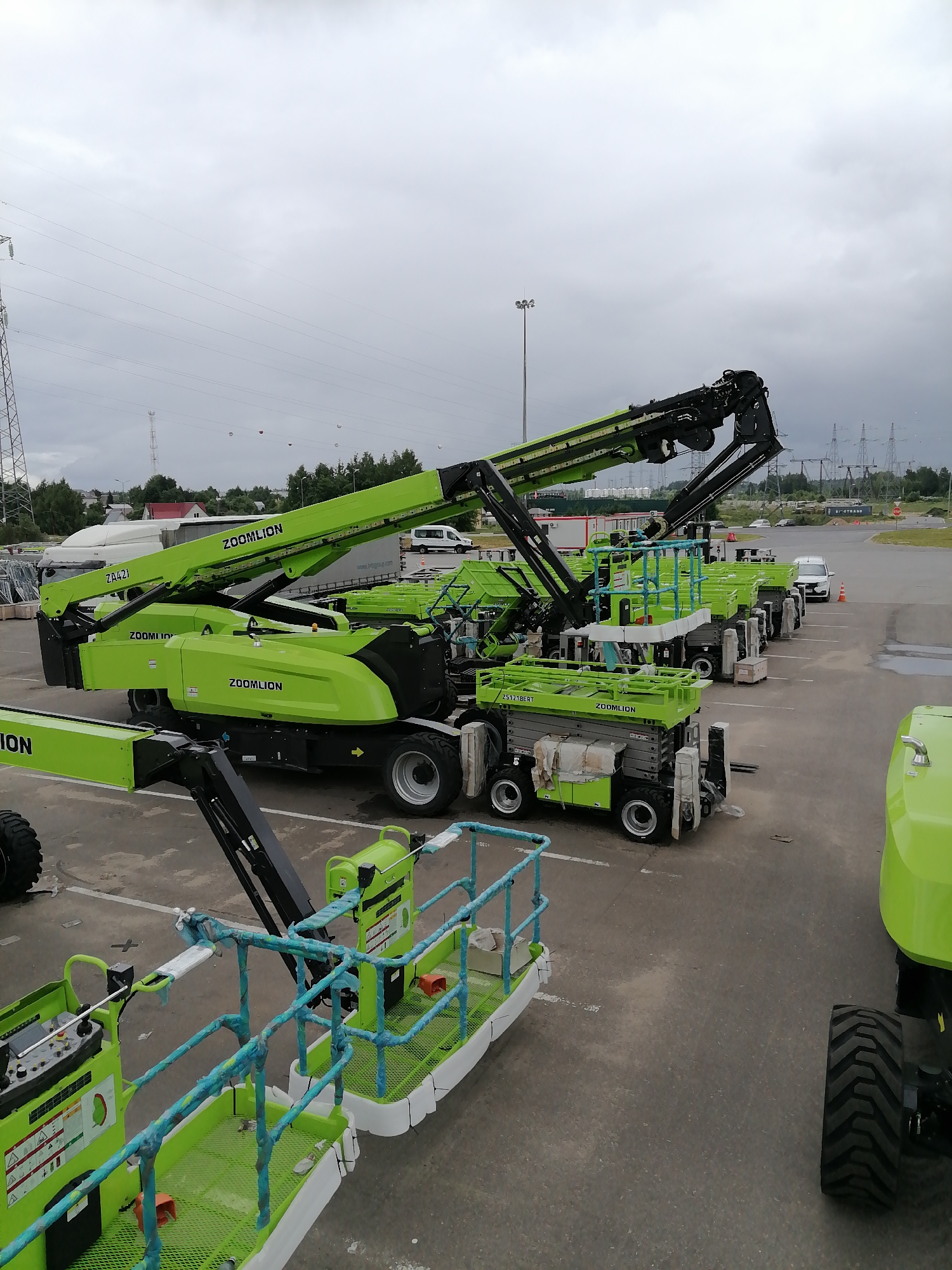 In Спас-Каменка, Russia, a fleet of vibrant green Zoomlion articulated boom lifts and scissor lifts are parked on a paved lot. The lifts are arranged in rows, with some of the boom lifts extended. The sky is overcast, and power lines and pylons are visible in the background, suggesting an industrial or semi-urban setting. The overall scene depicts a storage or staging area for construction equipment.