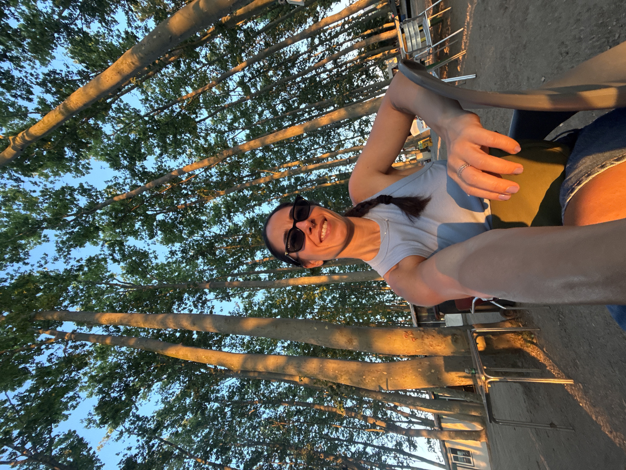 A woman with dark hair braided and wearing sunglasses sits in a chair outdoors, holding a phone to take a selfie. She is wearing a light blue tank top and denim shorts. Behind her, tall trees with sunlight filtering through them create a dappled effect. In the background, there are picnic tables and a building. The scene appears to be a park or outdoor dining area in Santa Cristina d'Aro, Spain.