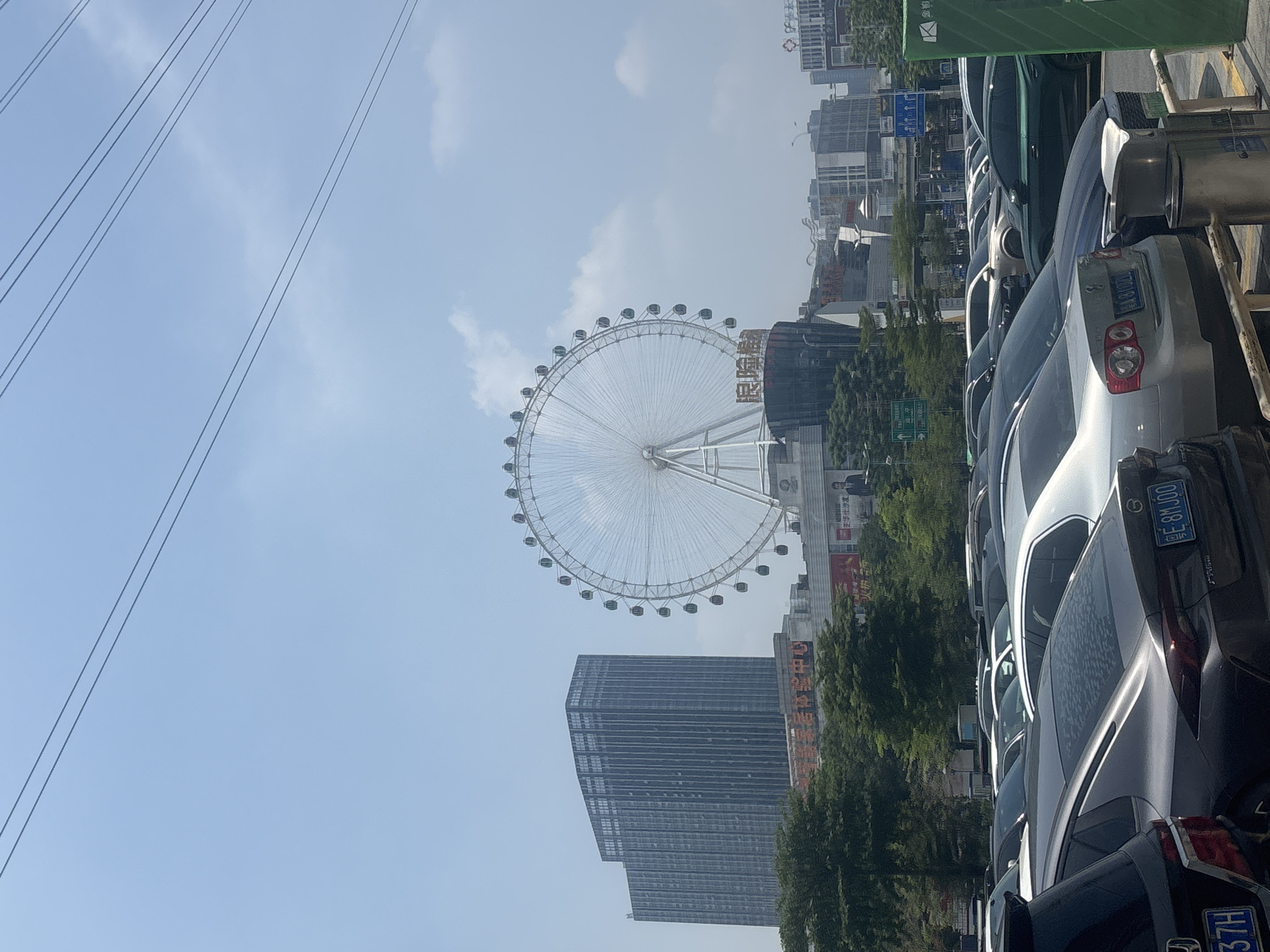 The image shows a daytime exterior view of a Ferris wheel in Nanhai District, China. The Ferris wheel is a large, white structure with numerous colorful gondolas. It is situated behind a parking lot filled with various cars. To the left of the Ferris wheel, a tall, modern, blue-tinted skyscraper dominates the skyline. In the background, other buildings and structures are visible, suggesting an urban environment. The sky is clear with a few scattered clouds. The scene appears to be a bustling city area, likely a commercial or entertainment district, given the presence of the Ferris wheel and the surrounding buildings. The focus is on the urban landscape with the Ferris wheel as a prominent landmark.