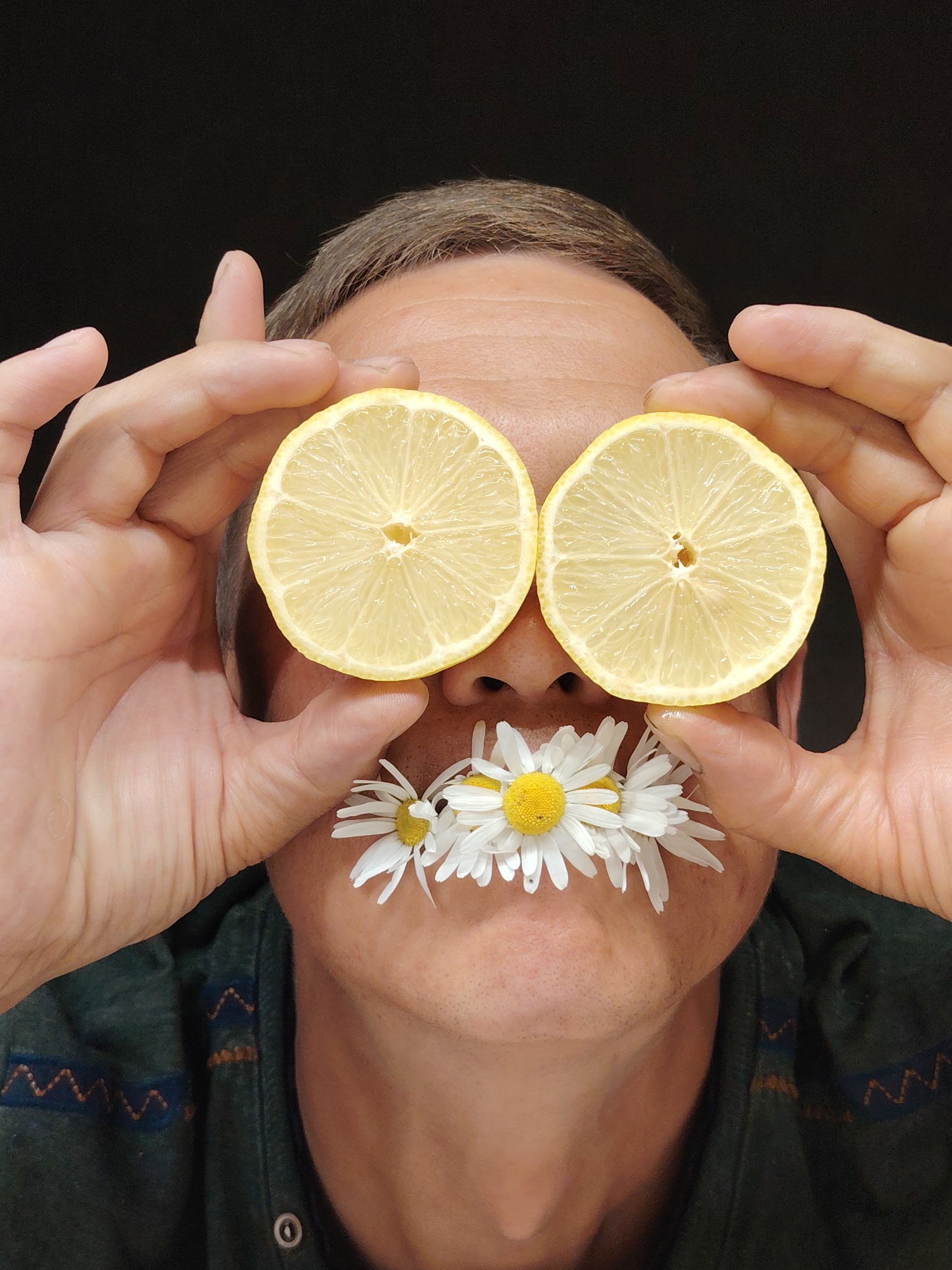 A close-up, head-and-shoulders shot shows a person against a dark, out-of-focus background. The person's head is tilted slightly upwards, and they are holding two slices of lemon to their eyes, with the fleshy side facing outwards. Their hands are positioned on either side of their face, with fingers gently curved. In place of the person's mouth, there are several white daisies with yellow centers arranged in a line. The person is wearing a dark green collared shirt with a patterned trim. The overall impression is playful and whimsical. The image's location in Chaikovsky, Russia, is not evident from the visual details provided.