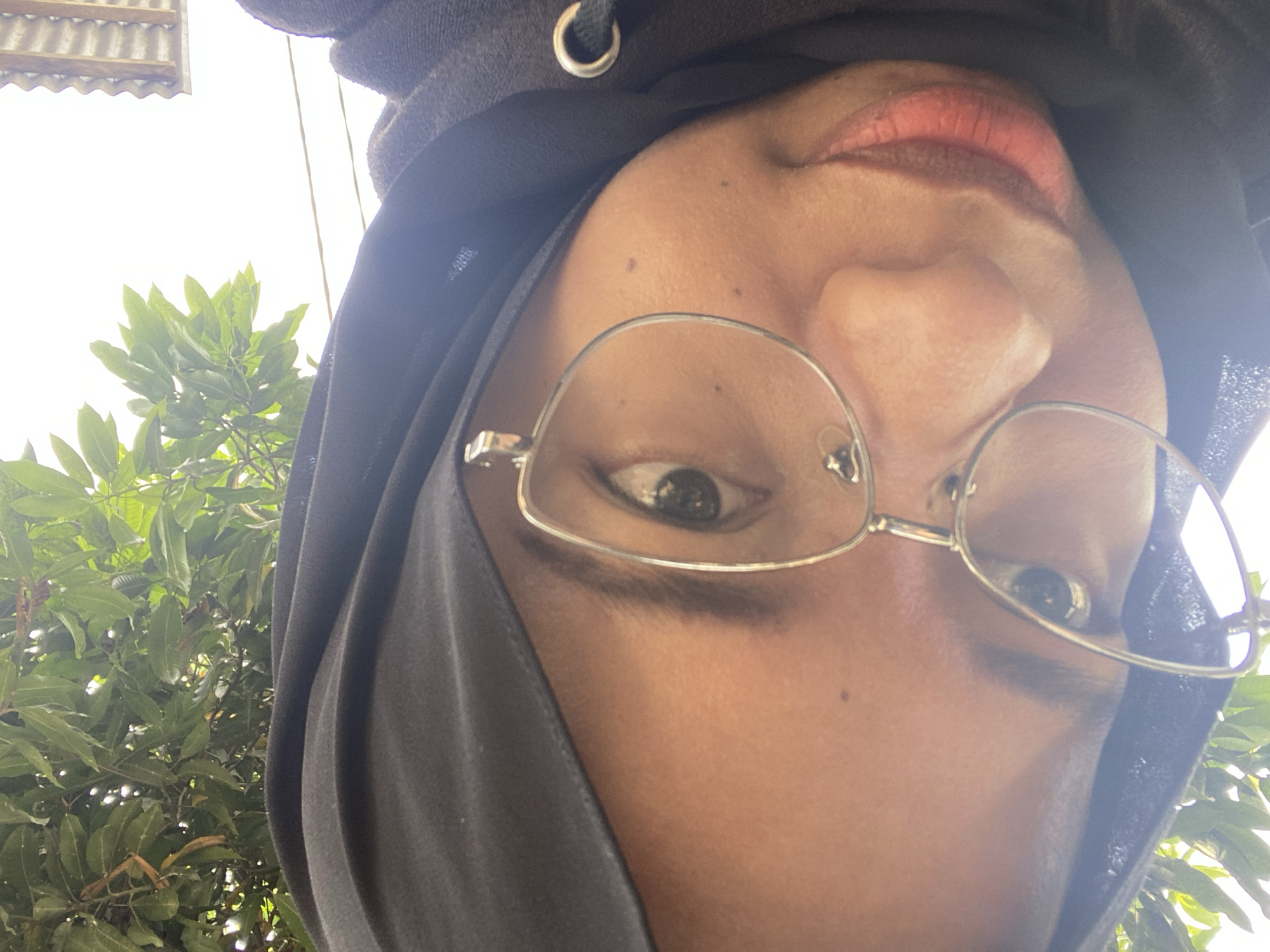 A close-up, low-angle selfie shows a young woman wearing a black hijab and clear-framed glasses. She is outdoors, with blurred green foliage visible at the top of the frame, suggesting a natural environment. Her head is tilted, and her face is partially illuminated by bright sunlight. The background is out of focus, with some hints of a roof structure and power lines visible, indicating an urban or semi-urban setting, potentially in Sekar Mawar, Indonesia. The overall impression is a casual, candid moment captured by the subject herself.