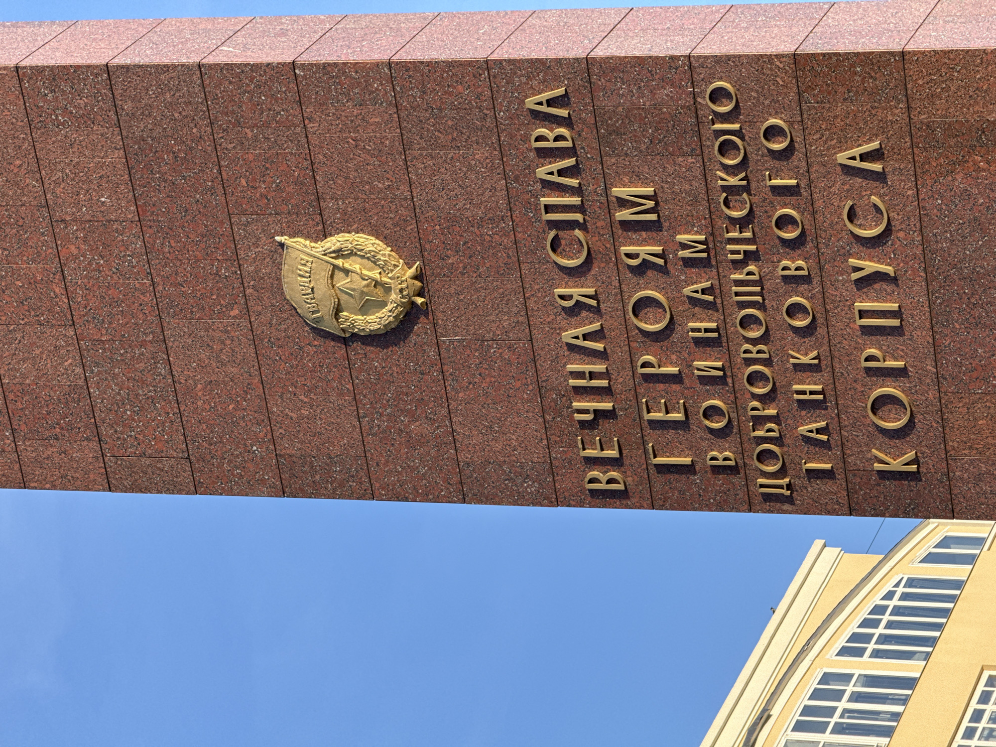 The image presents a close-up view of the Monument to the Ural Volunteer Tank Corps, located in Perm, Russia. The central subject is a tall, rectangular stele constructed from polished, reddish-brown granite tiles.
Affixed to the upper part of the granite is a golden metallic emblem. This emblem features a stylized flag inscribed with the Cyrillic word "ГВАРДИЯ" (Guard), positioned above a five-pointed star containing a hammer and sickle, all enclosed within a laurel wreath. Below the emblem, three-dimensional golden Cyrillic letters are mounted on the granite surface, spelling out: "ВЕЧНАЯ СЛАВА ГЕРОЯМ ВОИНАМ ДОБРОВОЛЬЧЕСКОГО ТАНКОВОГО КОРПУСА" (Eternal Glory to the Heroes, Soldiers of the Volunteer Tank Corps).
The background consists of a clear, brilliant blue sky, occupying the upper-left quadrant of the frame. A portion of a multi-story building is visible in the lower-left. This building features light-colored, possibly yellow or beige, walls and multiple rectangular windows with white frames, some arranged in an arched or stepped configuration along its upper facade. No individuals or specific actions are depicted within the scene. The overall lighting suggests bright daylight conditions.