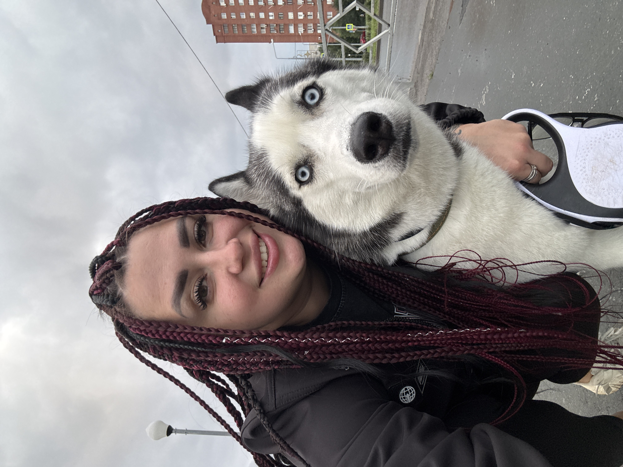 A selfie shows a woman with long, dark red box braids and a husky dog. The woman is smiling and wearing a black jacket. The dog has striking blue eyes and is looking at the camera. They are standing outside on a cloudy day, with a multi-story brick building in the background, characteristic of Tolyatti, Russia. There are streetlights and a metal fence visible in the background.