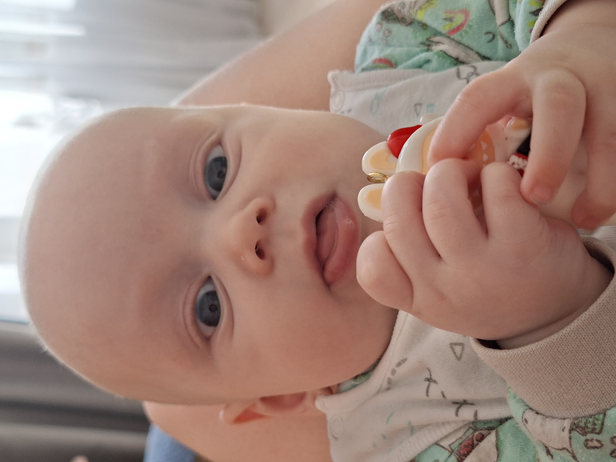A close-up shot focuses on a baby with fair skin and light blue eyes, lying on their back. The baby is wearing a pajama top with light green and white patterns and a beige cuff. The baby is holding a toy with a white ring, a red bead, and a small figure inside. The baby's tongue is slightly visible, protruding between their lips.