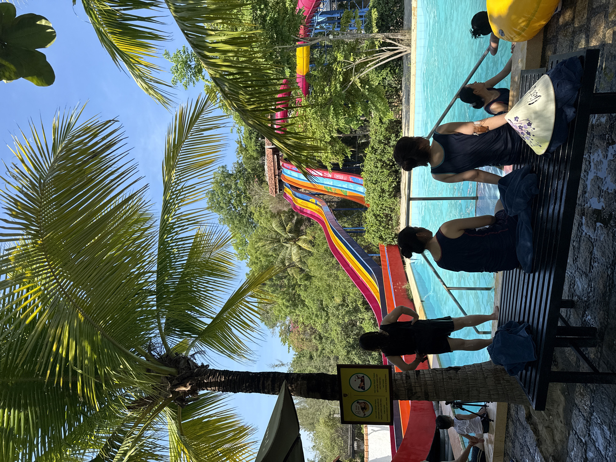 The image shows an outdoor scene in Tay Nha Trang Ward, Vietnam, featuring a water park. In the center-right of the image, a group of women stands next to a pool. One woman is reaching into the water. Near the women, there is a bench and a straw hat. Behind them, a colorful water slide is visible. Palm trees frame the left side of the image, partially obscuring the sky. The foreground includes a sign with Vietnamese text. A smaller water slide is visible in the background.