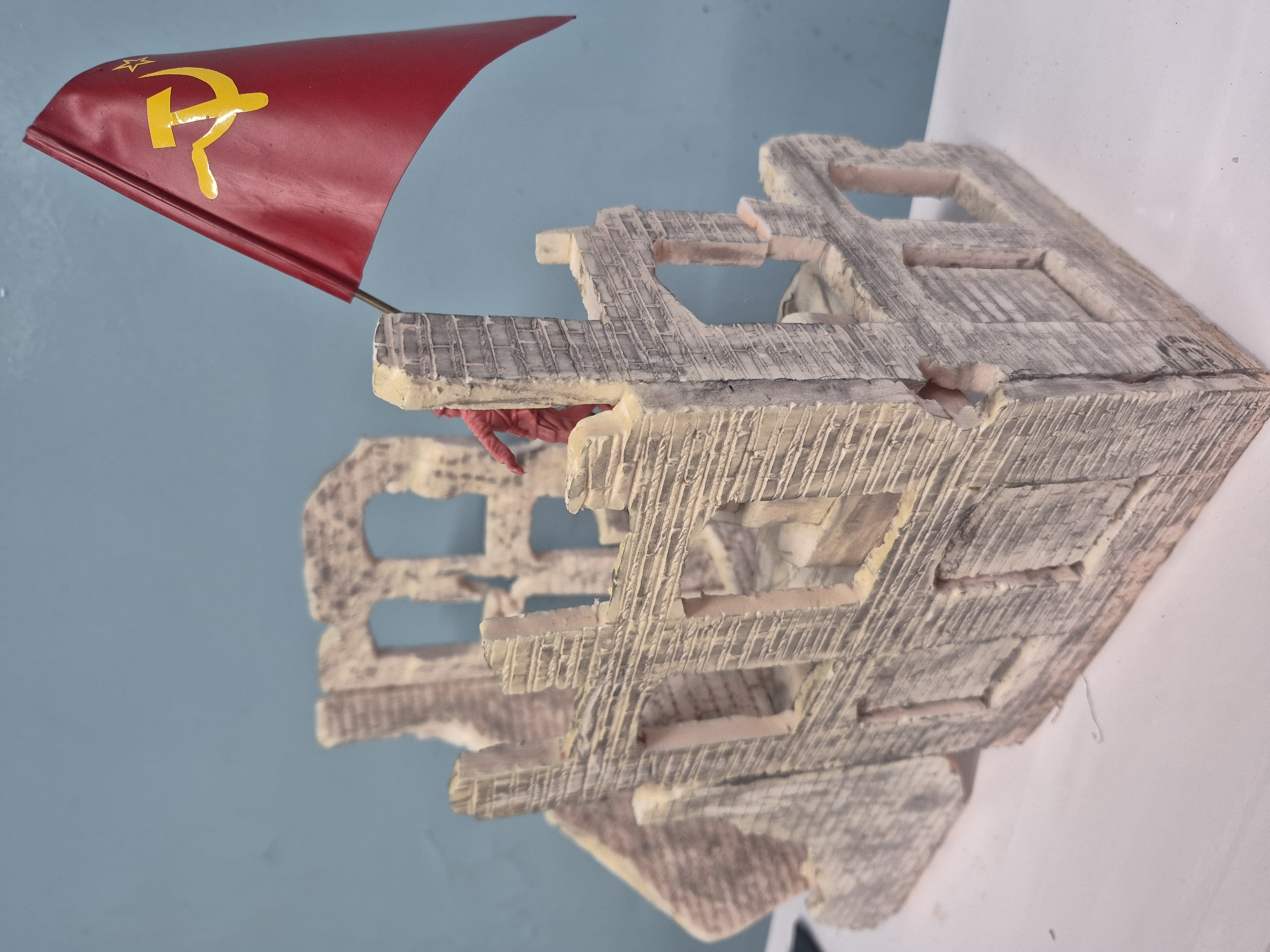 A low-angle, close-up shot shows a model of a ruined building, likely depicting a scene of conflict or destruction. Atop the building, a red flag bearing the hammer and sickle symbol of the Soviet Union is affixed. The model's surface appears textured, possibly to simulate damage or aging. The background consists of a wall painted in a muted blue color on the left and white on the right. The scene is illuminated by an even light source, highlighting the model's detail and the flag's vibrant color. There are no indications as to a city location such as Chaikovsky, Russia.