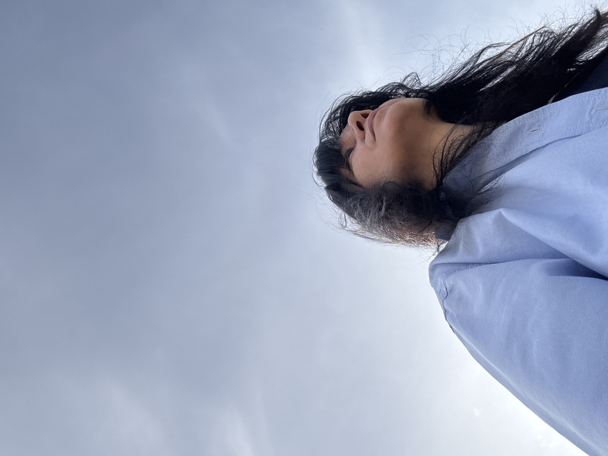 A low-angle photograph shows a person lying with their face tilted up towards the sky. The person has dark hair and is wearing a light blue button-down shirt. The background is a cloudy sky. It is not possible to determine the location as Chaikovsky, Russia from the image.