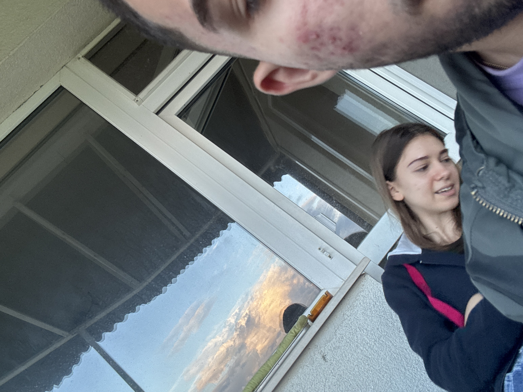 An unconventional close-up shot captures two individuals and a window reflecting a vibrant, cloud-filled sky, set in Blagoevgrad, Bulgaria. In the foreground, the lower part of a person's face, including the chin, jawline with stubble, and what appears to be acne or skin irritation, is visible upside down and extremely close to the camera. This person wears a dark green/grey jacket over a purple garment.
In the mid-ground, to the right, a young woman with brown hair is partially visible, looking towards the left with a slightly open mouth, suggesting she is speaking or smiling. She is dressed in a dark blue/black collared top with white trim and a prominent pink/red strap or ribbon crossing her chest.
A large window with light-colored frames dominates the left and center of the image. The glass reflects a colorful sky, displaying clouds with warm, golden, and orange hues, indicative of either sunrise or sunset. Inside the window, a scalloped-edged blind or shade is visible. A textured, light-colored wall is present on the left and bottom right. On the window frame or sill, a small orange object with a dark section and a green wrapped cable or pipe can be seen. No text is visible in the image.
