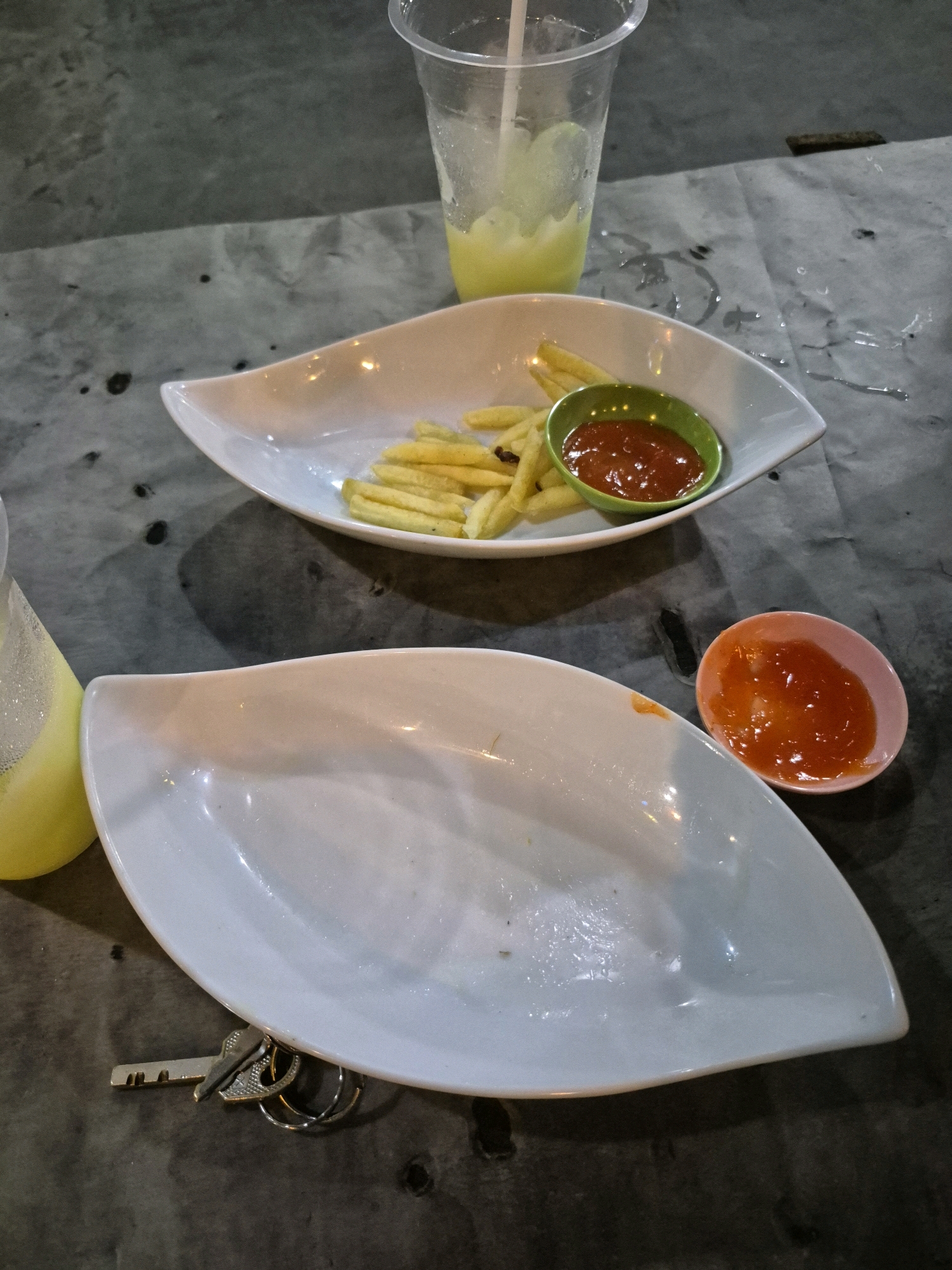 The image depicts an outdoor table setting at night, possibly at a street-side food stall or casual eatery, with a concrete floor that appears slightly damp or stained, indicative of an outdoor environment. Two white, leaf-shaped plates are present. The upper plate contains a small portion of french fries and a small green dish with red sauce, likely chili sauce or ketchup. The lower plate is empty, save for a small smear of sauce. Next to the lower plate, a small pink dish also holds red sauce. In the background, a clear plastic cup with a white straw holds a yellow beverage, possibly juice or iced tea. A set of keys with a silver keychain is visible beneath the edge of the lower plate, resting on the concrete. The lighting suggests artificial light sources, creating shadows on the table and ground. The overall scene suggests the remnants of a meal. Based on the prompt, this scene is located in Pabelan, Indonesia.