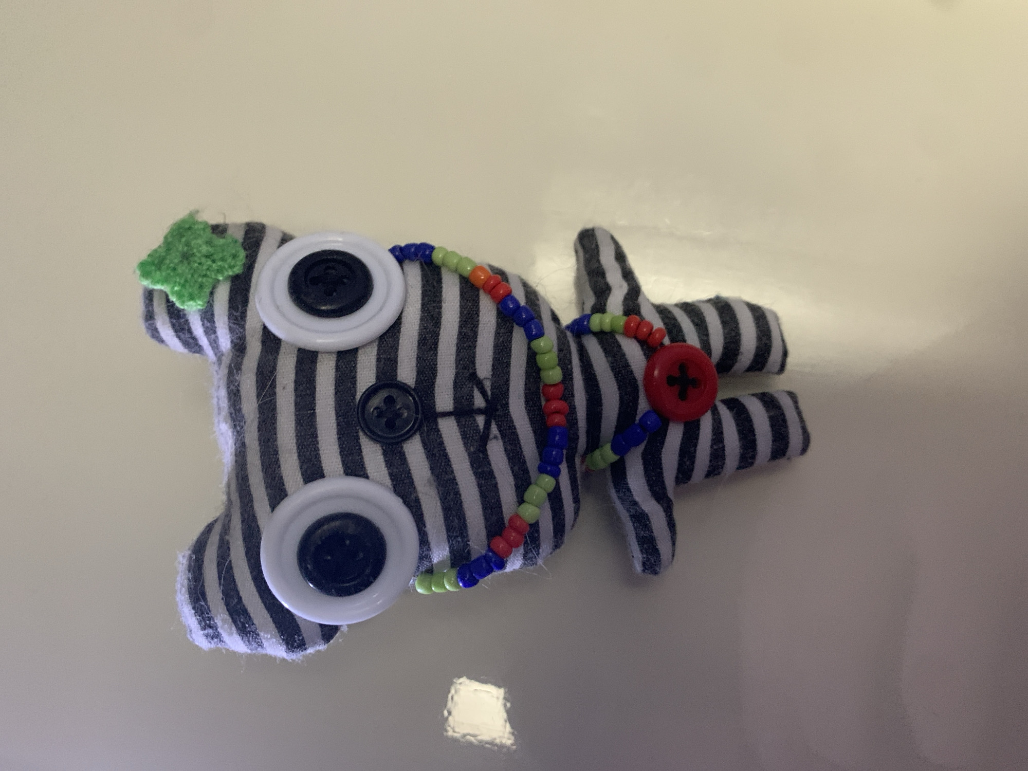 This media file depicts a handmade stuffed toy lying on a light-colored, reflective surface. The toy is primarily black and white striped, with four white button eyes, each with a black button in the center. One eye appears to be missing its black button. There is a small, green, textured fabric embellishment on the upper left side of the toy, possibly representing an ear or a decorative element. A beaded necklace, comprised of green, orange, red, and blue beads, is draped around the neck area of the toy. A red button with four black holes, also sewn on, is visible on the right side of the toy. The toy's body is somewhat flat, with the limbs extended outwards. The overall impression is of a whimsical, handcrafted item. The lighting suggests an indoor setting, possibly with artificial light, as evidenced by the bright reflections on the surface. There is no visible text in the image. The background is a plain, off-white color. The setting does not offer specific clues about the location, but the provided context mentions Severodvinsk, Russia.
