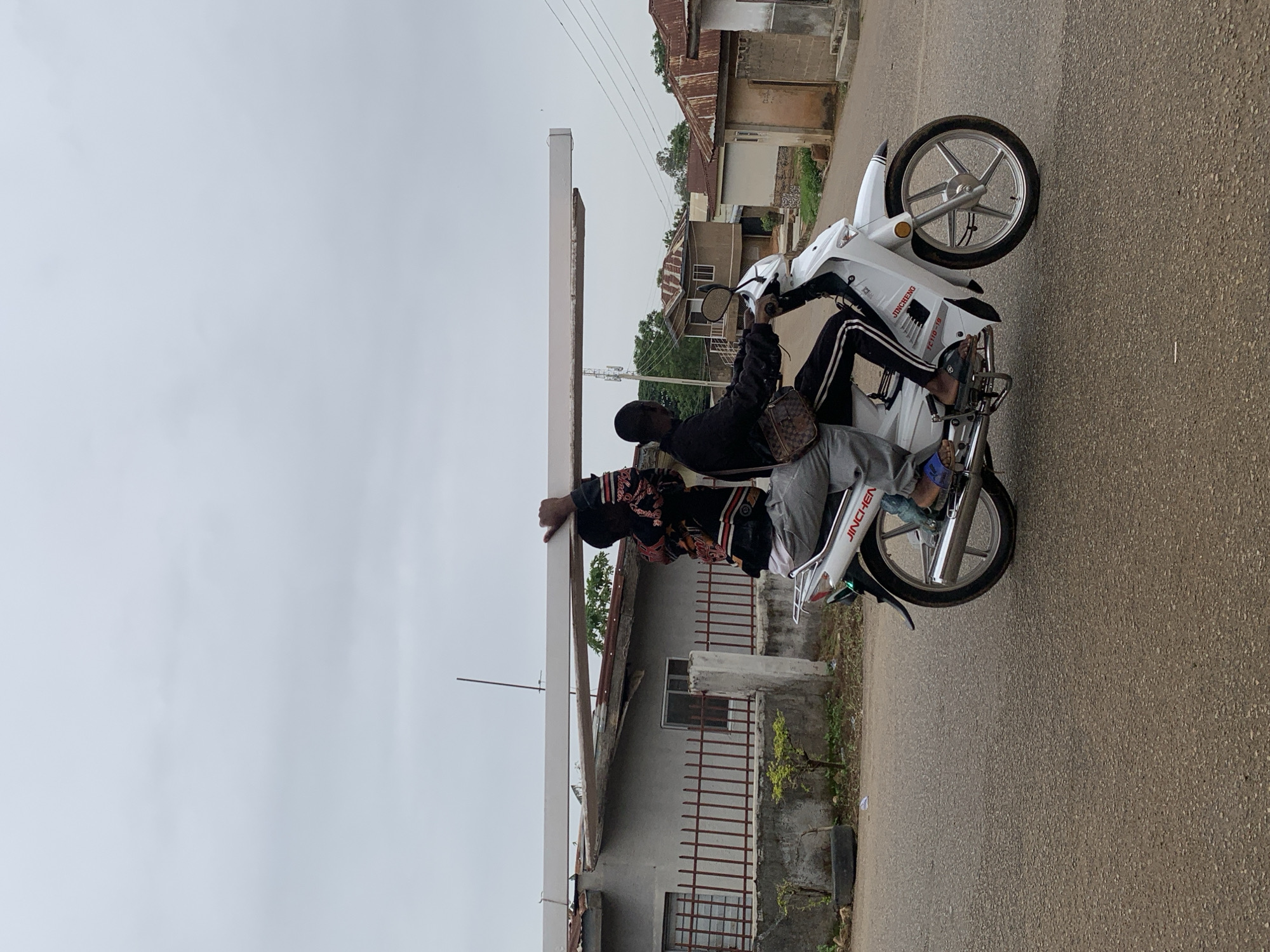 Two men are riding a white Jingcheng TC110-19 motorcycle on an asphalt road in Jos, Nigeria. The rider is wearing a black jacket and black pants with white stripes. The passenger behind him is wearing a colorful jacket and grey pants. The background shows buildings and an overcast sky.