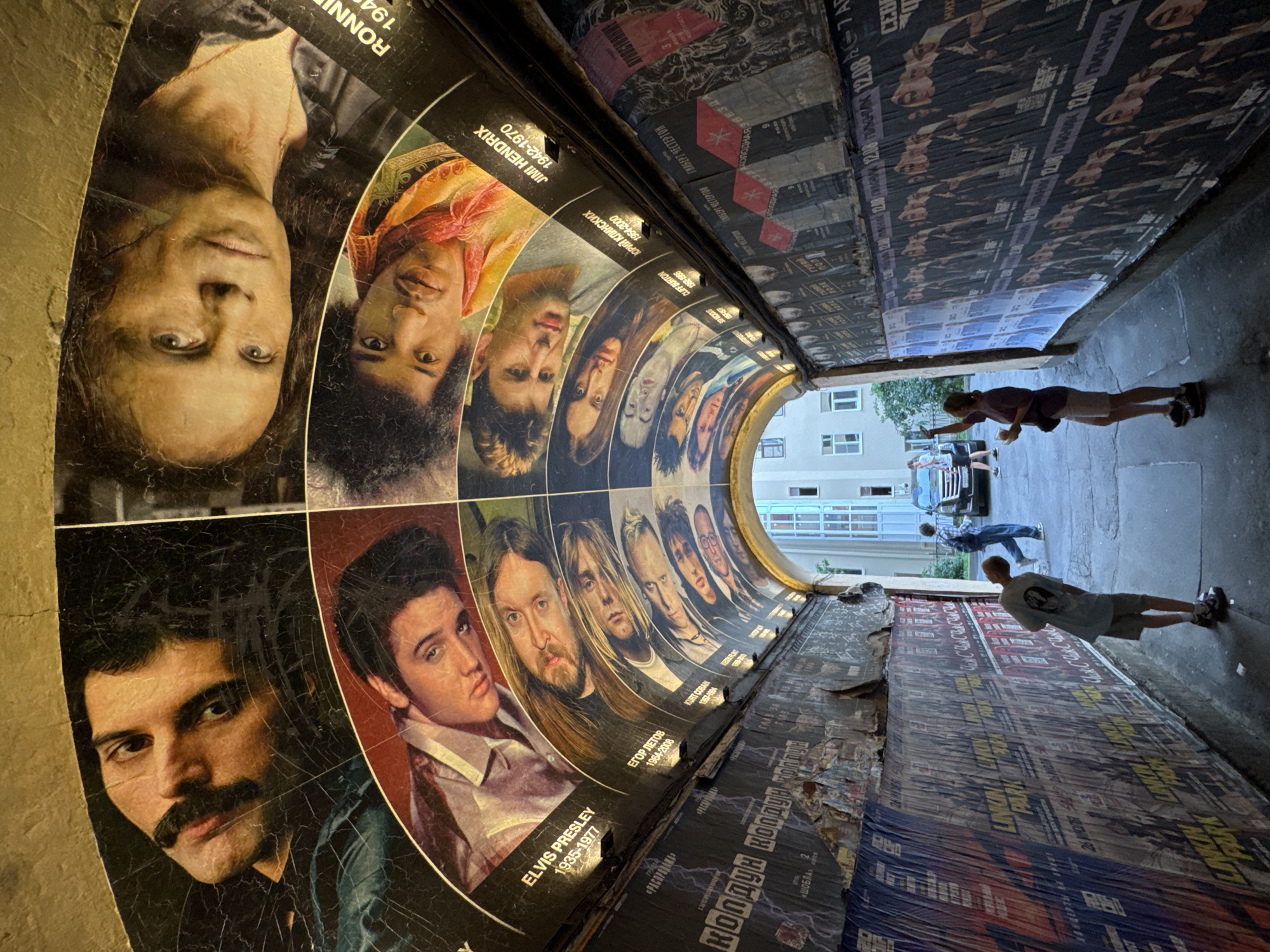 The image depicts a tunnel or archway in Saint Petersburg, Russia, decorated with large posters of musicians and other figures on its curved ceiling. The posters are of various musicians including Elvis Presley, Jimi Hendrix, and Kurt Cobain, as well as other figures like Freddie Mercury and Ronnie James Dio. Below these, on the walls of the tunnel, are more posters, some of which appear to be for concerts or events, with some featuring the band Linkin Park. Two individuals, a man and a woman, are visible in the tunnel. The man is wearing a light-colored t-shirt and shorts, and is looking at the posters on the wall. The woman is wearing a darker t-shirt and shorts, and is holding a drink while looking at her phone. A vehicle is parked in the background, and a building with windows is visible at the end of the tunnel. The overall condition of the posters suggests age, with some cracking and peeling.
