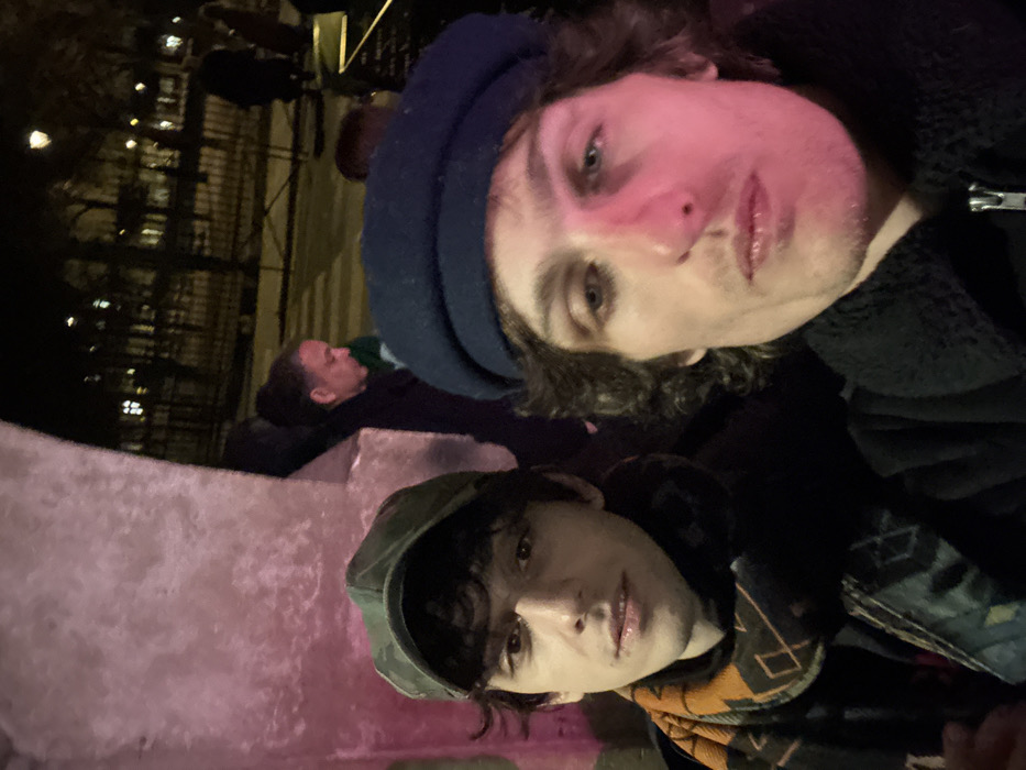 Two men pose closely for a selfie in low light. The first, wearing a dark blue beret and dark jacket, has fair skin, light eyes, and reddish cheeks. Below, the second man sports a green cap and a colorful patterned scarf. Behind them, a person is lying down on what appears to be a concrete ledge, with streetlights and dark buildings in the background. The image has a casual, candid feel.