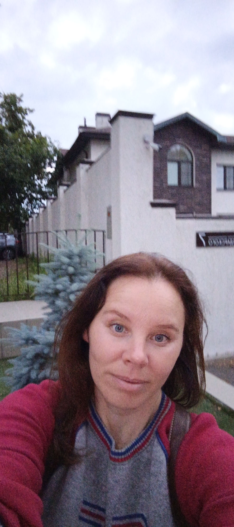 A woman with shoulder-length brown hair and blue eyes takes a selfie outdoors. She is wearing a red and grey sweater with blue and red trim around the collar. Behind her is a modern house with light-colored walls, a dark-tiled roof, and arched windows. A fence and a small, blue-green conifer tree are visible in front of the house. The sky is overcast. The location is identified as Votkinsk, Russia.