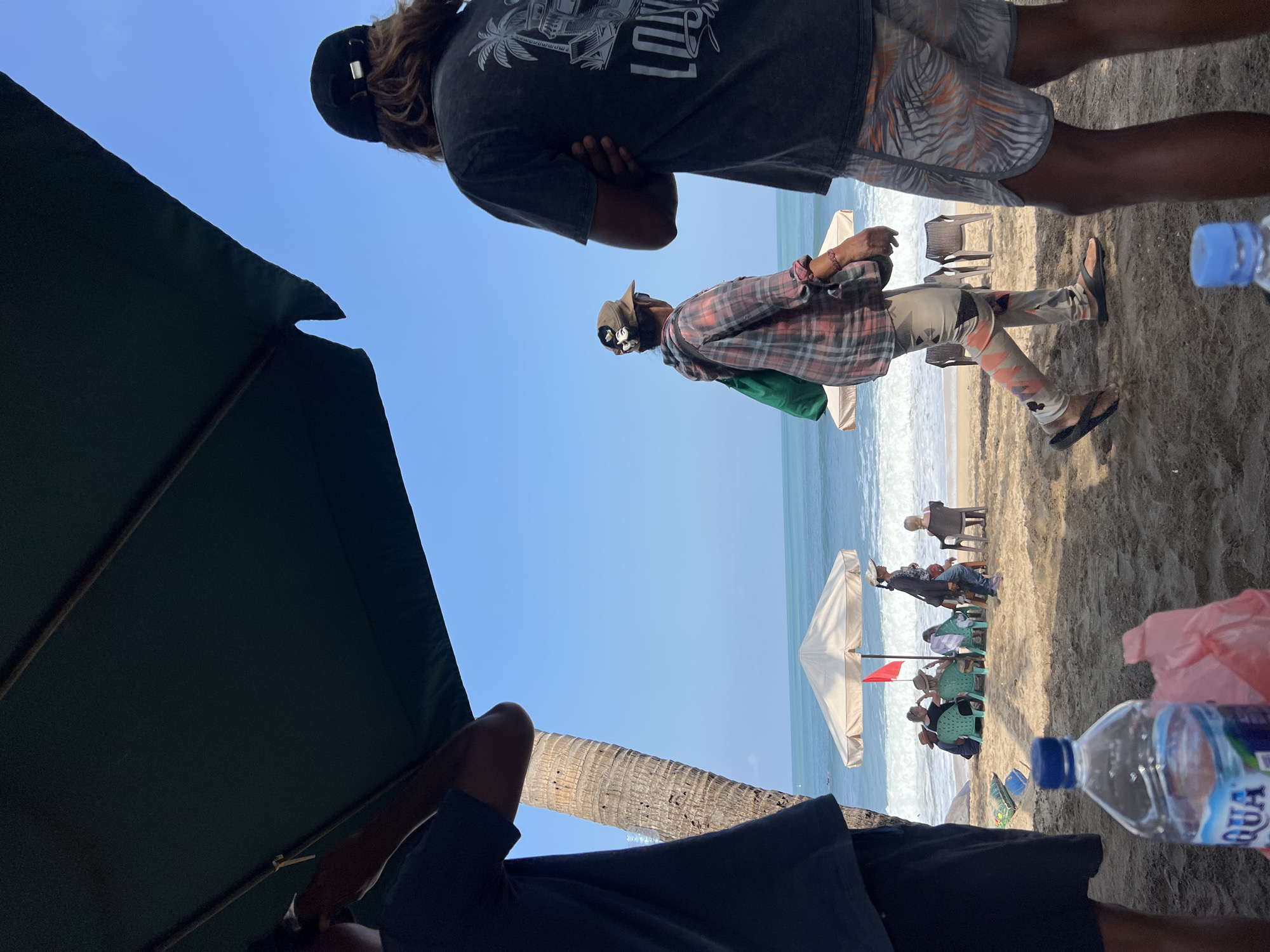 The image depicts a sunny day on Kuta Beach, Indonesia. In the foreground, a person wearing a black t-shirt and patterned shorts stands with their arms crossed, facing away from the camera. To their left, another person in a dark t-shirt is partially visible, also facing away. Between them, a large dark umbrella casts a shadow over part of the beach.
Further in the background, a person wearing a plaid shirt and patterned leggings walks with a green bag slung over their shoulder. They are walking towards the ocean, which is visible with moderate waves. Several beach umbrellas are set up, with people seated in chairs beneath them, enjoying the beach. One prominent umbrella is white with a red flag planted in front of it. A tall palm tree trunk is visible on the left side of the frame.
In the immediate foreground, a plastic bottle of "AQUA" mineral water is placed on the sand, alongside a crumpled pink plastic bag. The sand is a light brown color, and the sky is a clear, bright blue. The overall scene conveys a relaxed atmosphere typical of a beach resort.