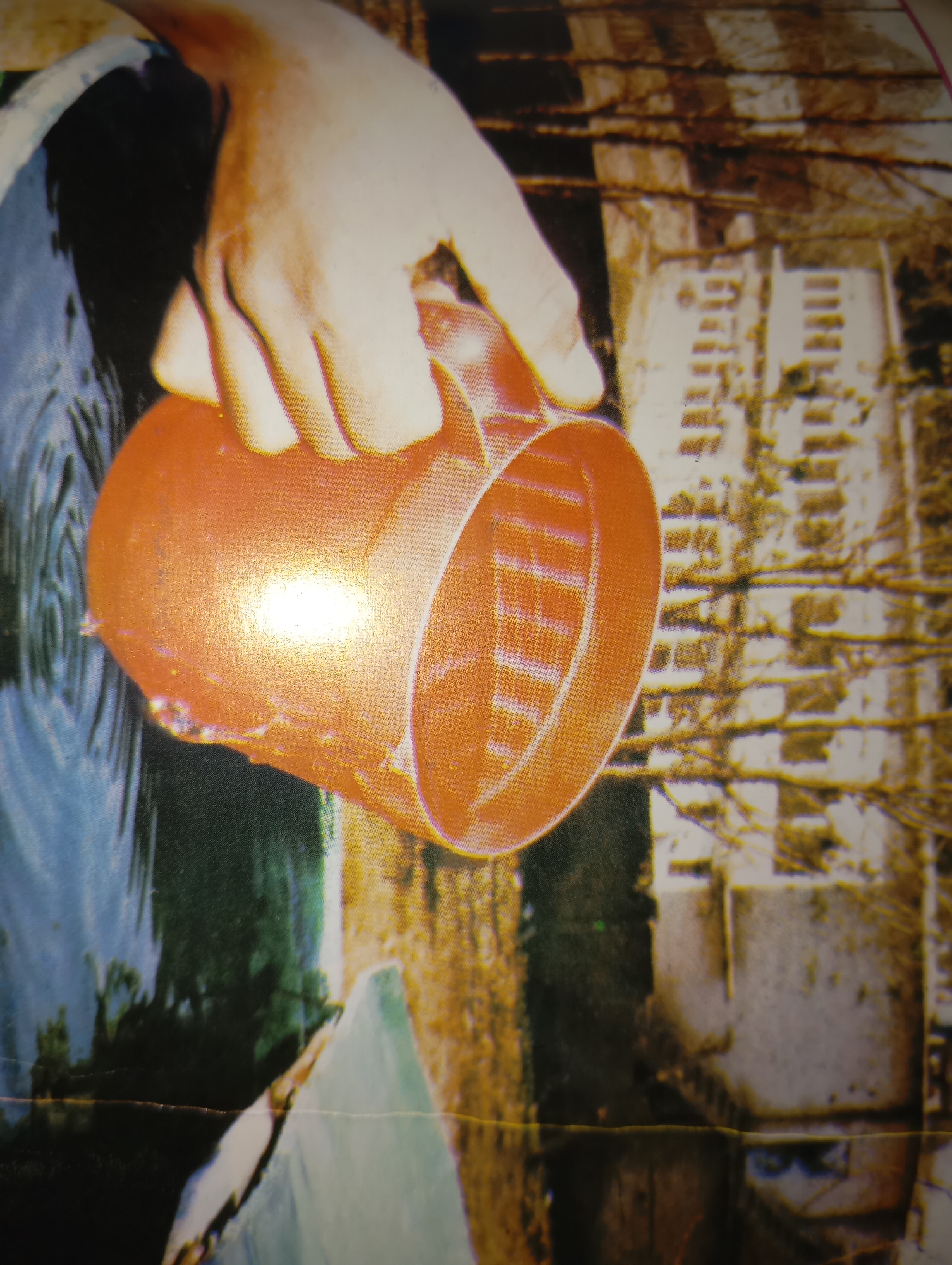 A close-up photograph captures a human hand holding an orange bucket over rippling water. The bucket appears to be made of plastic and is tilted, with its opening facing downwards, as if to scoop water. The hand is positioned above the bucket, with fingers gripping its rim. The water is a deep blue-green and reflects the surroundings. In the background, a blurred, indistinct structure is visible, appearing to be made of light-colored bricks or concrete blocks, with horizontal lines suggesting shelves or tiers. The overall scene is outdoors, with elements of natural water and man-made construction. The location is stated to be the city of Dangari, Pakistan.