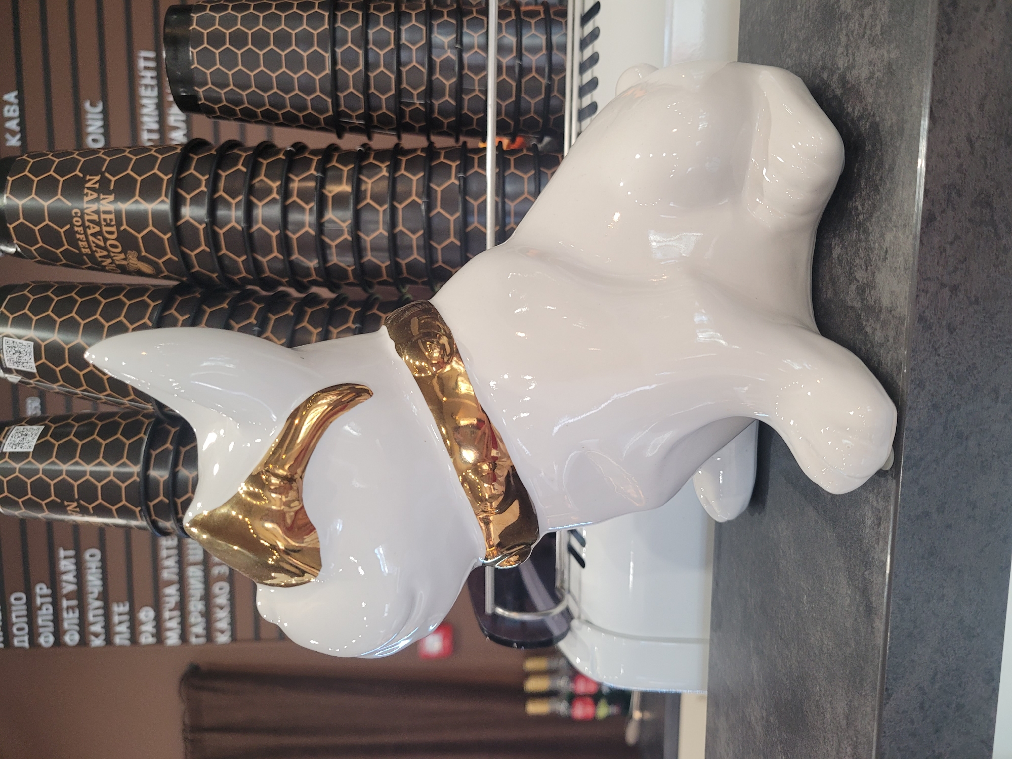 A white ceramic French bulldog figurine with gold accents sits on a dark grey countertop. The figurine is in the foreground, with its head turned to the left, wearing gold-rimmed glasses and a gold collar. Behind the figurine, stacks of coffee cups with a honeycomb pattern are visible, along with a portion of a white coffee machine. In the background, a brown wall displays a menu with Cyrillic text, indicating the location is a coffee shop. The menu lists items such as "КАПУЧІНО" (Cappuccino), "ЛАТЕ" (Latte), and "КАКАО" (Cocoa). The overall lighting suggests an indoor commercial setting. The scene is static, with no visible people or ongoing actions.