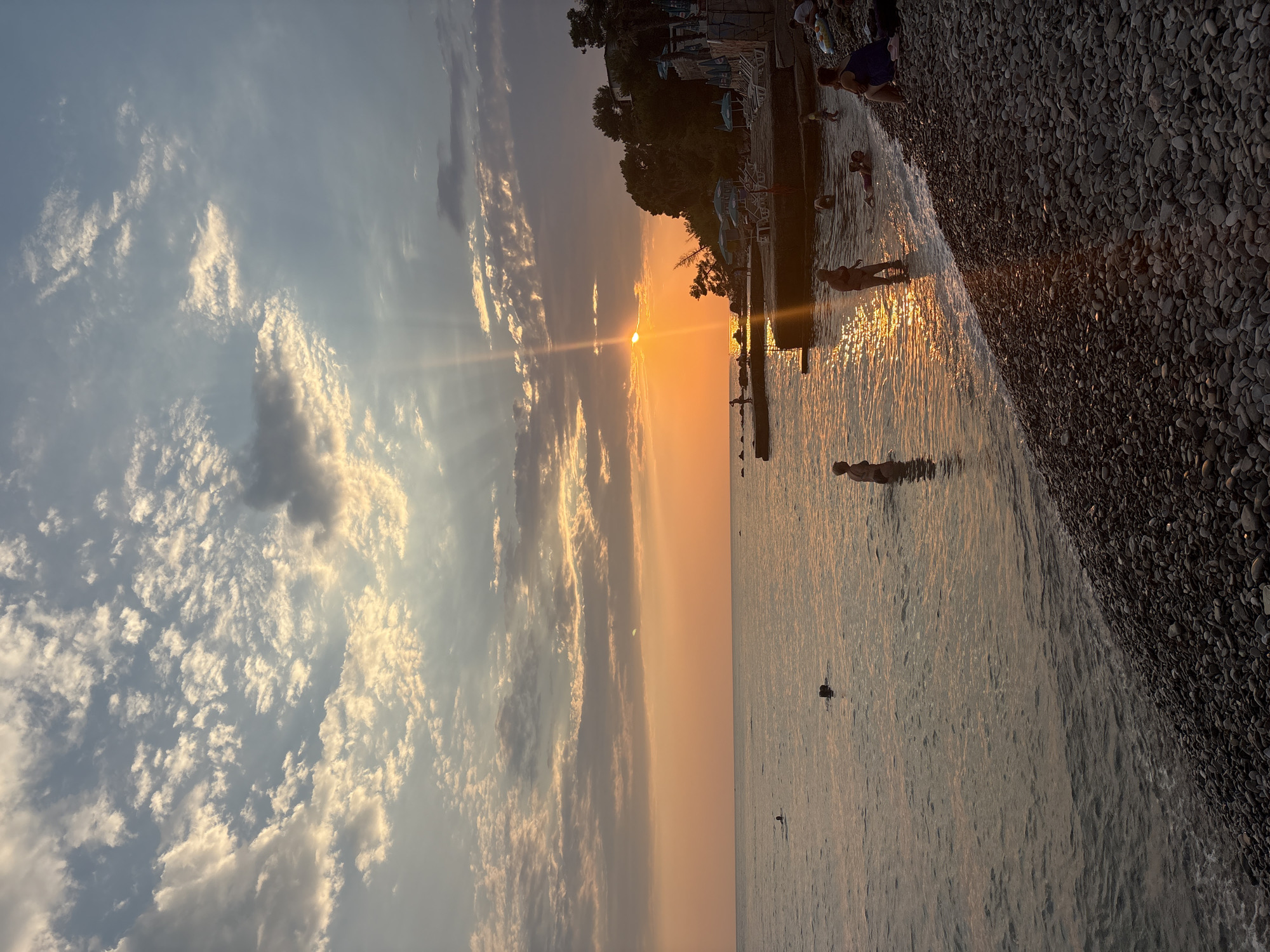 The image shows a beach scene at sunset in Bar, Montenegro. The sun is low in the sky, casting a warm orange glow over the water and clouds. The sky is filled with scattered clouds, some of which are illuminated by the setting sun. The sea is calm, with gentle waves lapping at the pebble beach. Several people are visible in the water and on the beach, some swimming, others relaxing. In the background, there are trees and buildings lining the coast. The overall atmosphere is peaceful and serene.