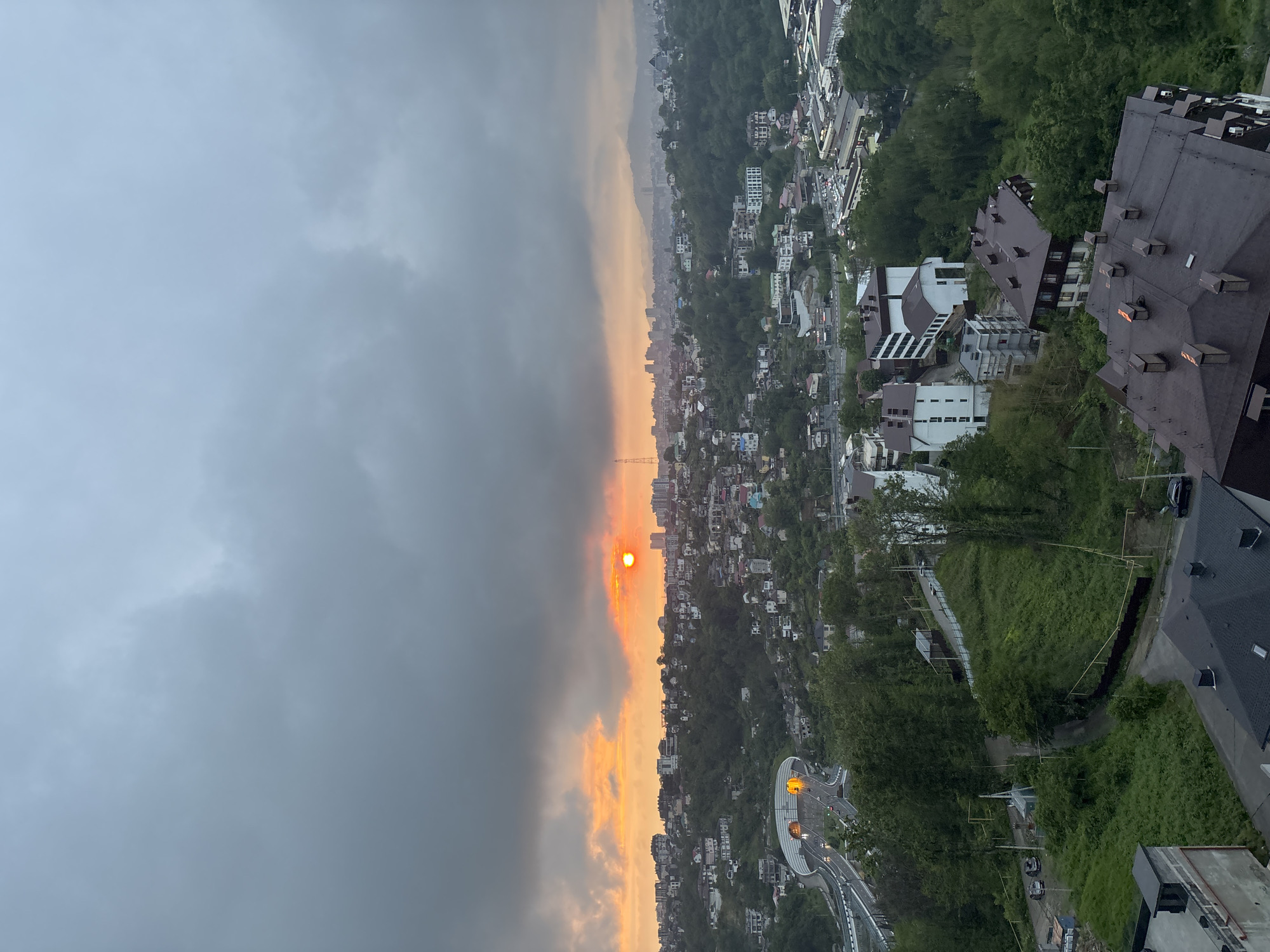 The image shows an aerial view of a cityscape during sunset or sunrise, possibly in Sochi, Russia. The sky on the left is dominated by a dark, cloudy mass, while the right side exhibits a bright orange-yellow gradient where the sun is positioned. The city is densely populated with buildings intermingled with green vegetation, suggesting a mix of urban and natural elements. In the foreground, some building rooftops are visible, offering a closer perspective on the urban landscape. A road with visible traffic lights and cars cuts through the city, indicating transportation infrastructure.