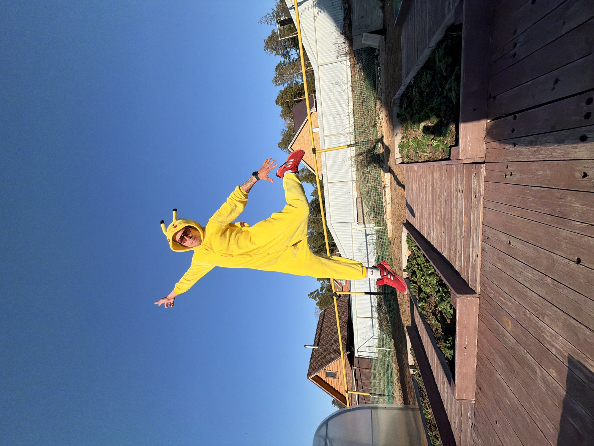 A man in a yellow Pikachu costume and red sneakers is performing a yoga pose with one leg extended upward, outdoors on a wooden deck next to a building with a white fence. He is in the city of Chaikovsky, Russia. The sky is clear and blue.