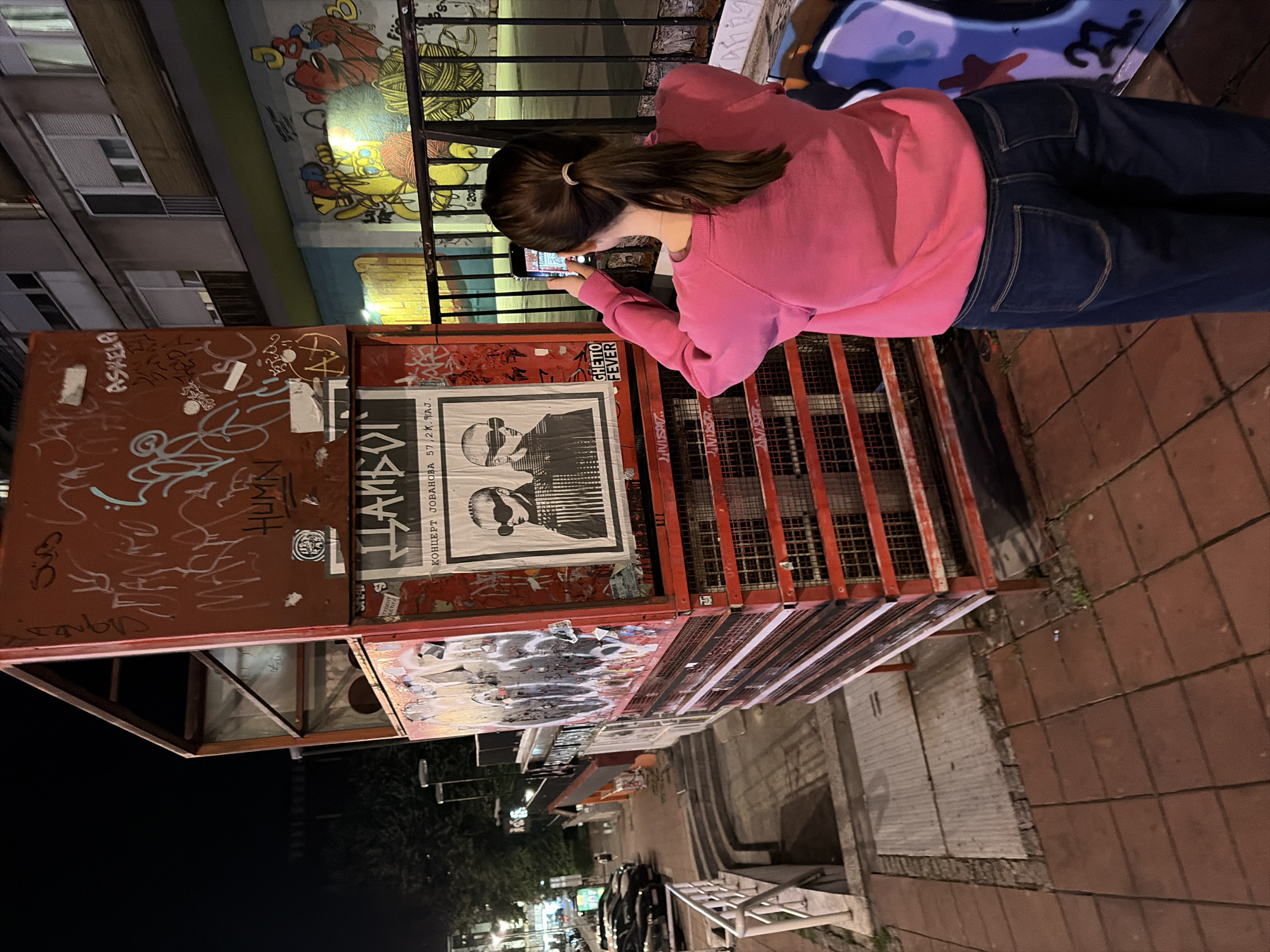 A person with long brown hair in a ponytail, wearing a bright pink sweatshirt and dark blue jeans, stands facing away from the camera, holding a smartphone to take a photograph of a tall, red, metal structure covered in graffiti and posters. The structure appears to be a kiosk or some kind of street furniture. One prominent poster on the structure features a black and white image of two people wearing large, dark sunglasses. Below this poster, the words "GHETTO FEVER" are visible.
The scene is outdoors at night, with artificial lighting illuminating the area. In the background, a multi-story building with windows and balconies is visible, as well as a brightly colored mural. To the left, a street with parked cars and a sidewalk with stairs leading down can be seen. The ground is paved with reddish-brown tiles and some grey concrete.
This scene is located in the Savamala neighborhood of Belgrade, Serbia, specifically near the Beton Hala, a popular waterfront area known for its nightlife and cultural venues. The graffiti and street art are characteristic of the urban landscape in this part of the city.