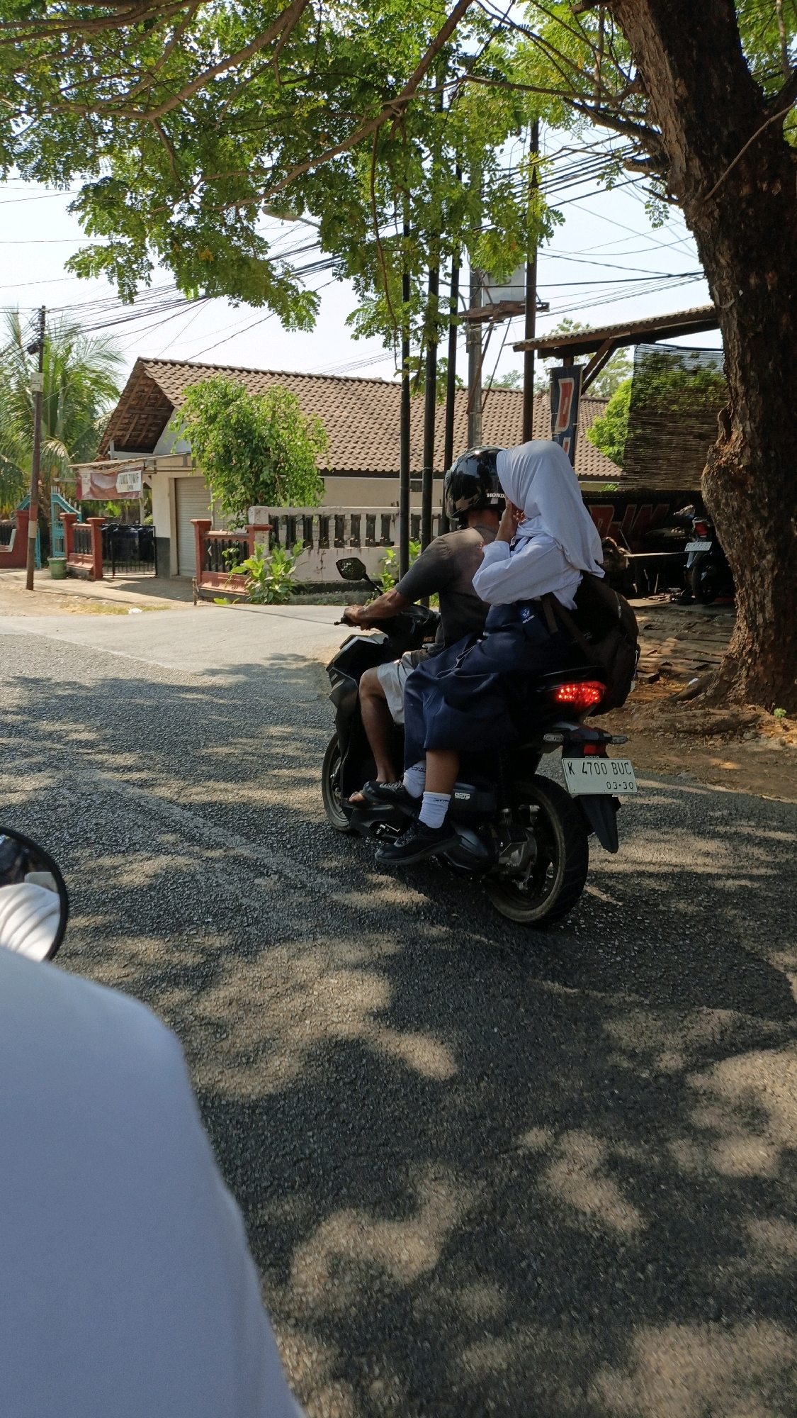 In Bandengan, Indonesia, on a sunny day, two individuals are riding a motorcycle on a paved road. The driver, wearing a dark t-shirt and a helmet, is operating the vehicle. Behind them, a younger person, dressed in a school uniform including a white hijab and a navy blue skirt, sits on the back. They are wearing white socks and black shoes, and carrying a backpack. The motorcycle has a visible license plate that reads "K 4700 BUC" with a date stamp "03-20". The environment includes trees with dappled sunlight casting shadows on the road, a building with a tiled roof, and power lines overhead. The perspective suggests the image is taken from another vehicle or person following behind.