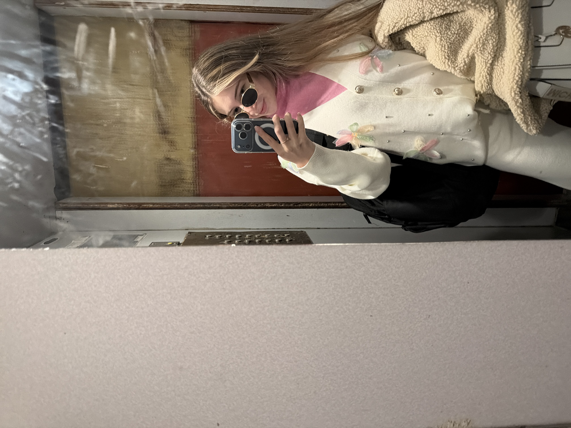 The image shows a woman taking a selfie in what appears to be an elevator in Plovdiv, Bulgaria. She is wearing sunglasses, a pink turtleneck, a white sweater with flower appliques, and a light brown sherpa jacket. A black bag hangs on her shoulder. The elevator has a metallic control panel and a partially visible background with yellow and red colors, which suggests a design element.