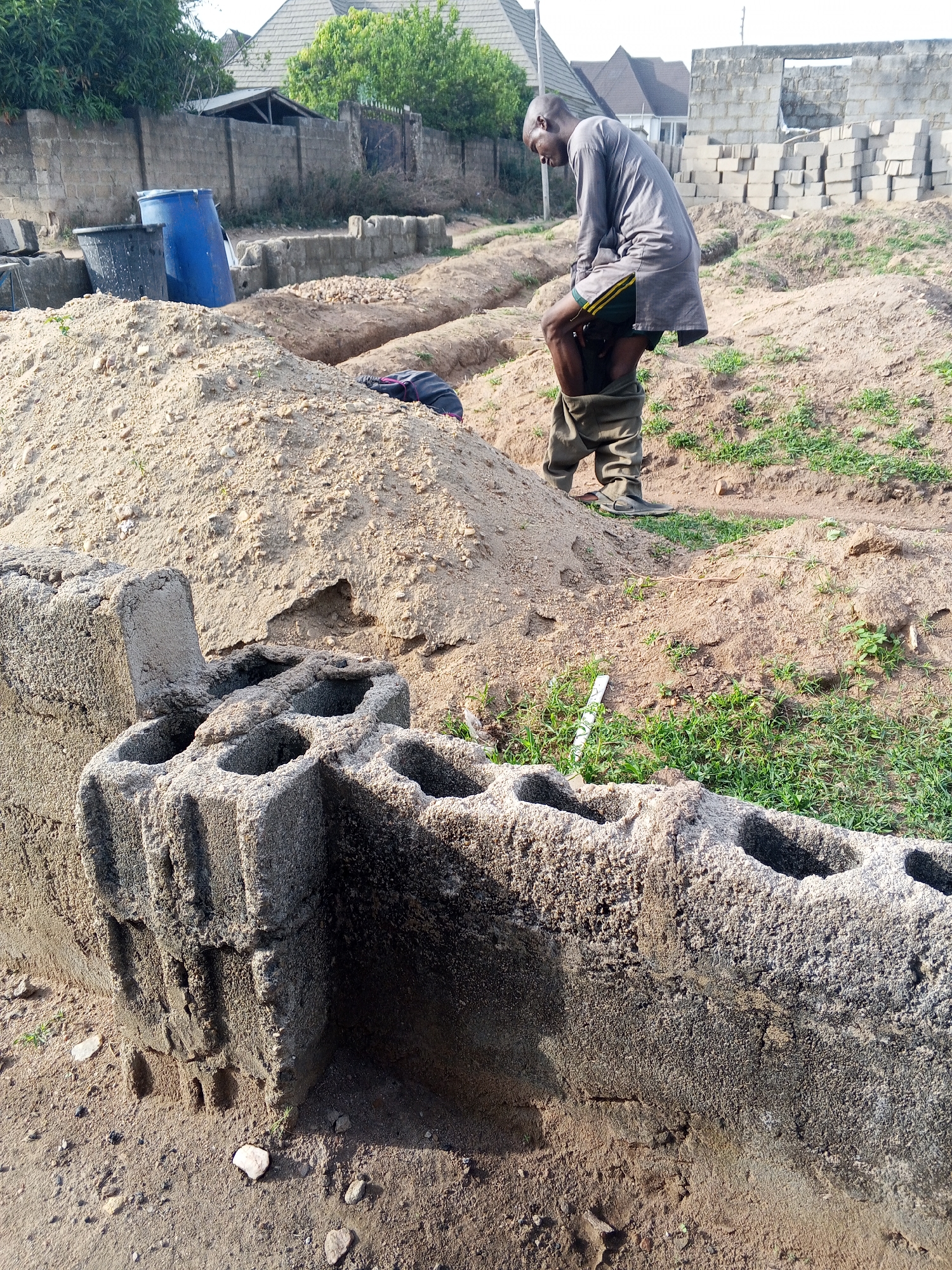 The image captures an outdoor scene, likely a construction site or an area under development, in Zango-ndaji, Nigeria, during bright daylight. The central human subject is a man, seen from the side, wearing a grey long-sleeved tunic and green shorts. He is actively adjusting or pulling up a pair of khaki or olive-green trousers that are gathered around his calves, with his head bowed down towards them.
The ground is predominantly sandy, featuring several mounds of loose sand and excavated trenches, characteristic of foundation work. In the immediate foreground, a low, partially built structure or foundation wall made of rough, hollow concrete blocks stretches across the frame. These blocks show signs of wear and a coarse texture. Behind the man, patches of green grass grow amidst the sandy terrain.
In the background, a large blue plastic barrel and a black container are visible next to a concrete block wall. Further back, stacks of concrete blocks are neatly piled, ready for construction. Beyond these elements, several buildings with roofs are discernable, some appearing more complete than others, under what seems to be a clear sky. No visible text is present in the image.