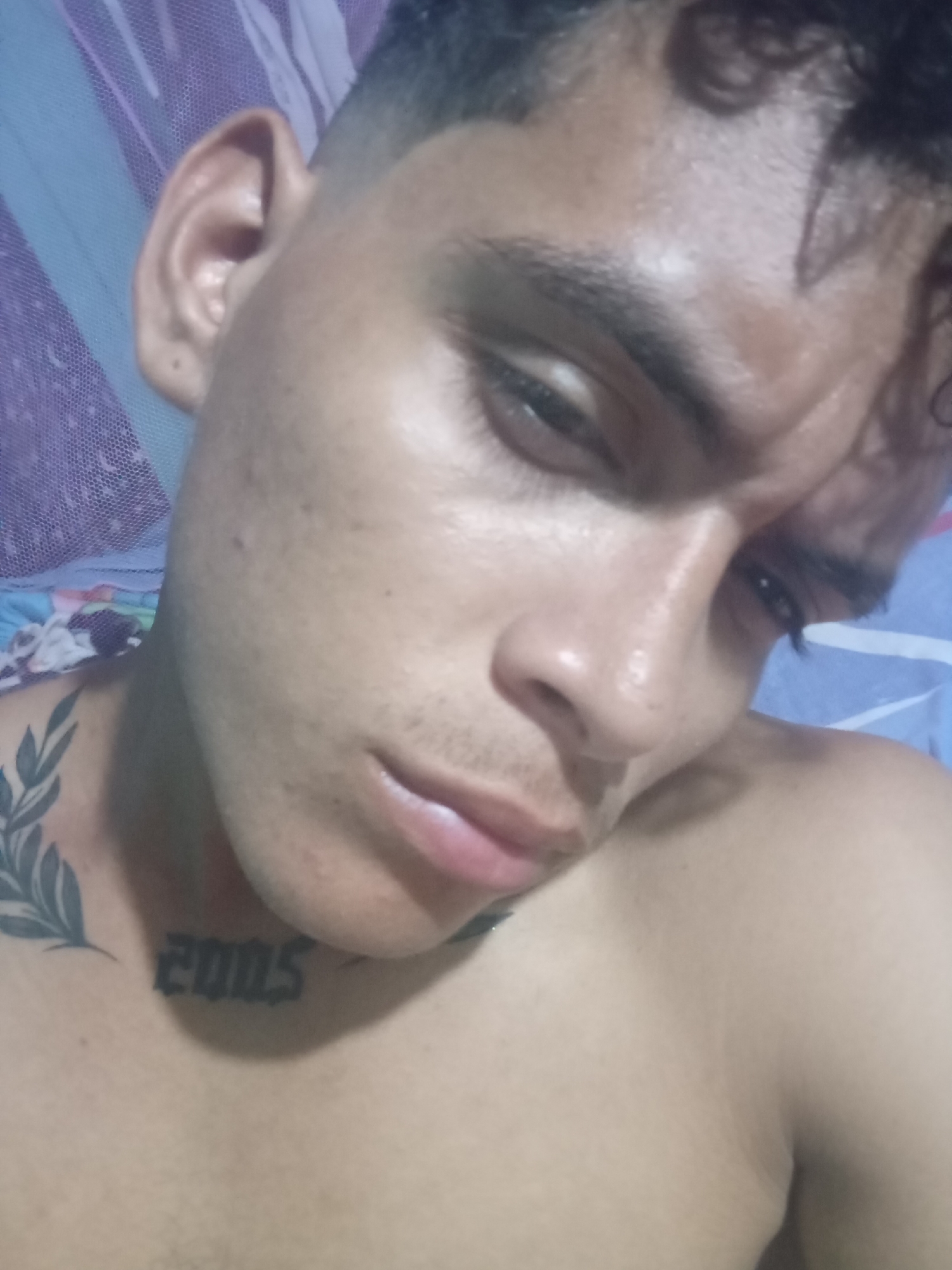 A close-up, angled shot of a young man's face and upper torso. He has short, dark hair and light brown skin. His expression is subdued, with his eyes downcast. A tattoo of laurel branches is visible on his left collarbone, and the numbers "2005" are tattooed on his neck. The background includes a blue and purple patterned fabric, possibly a mosquito net or bedding. The setting is unknown, but the prompt suggests it may be in Altagracia, Venezuela.