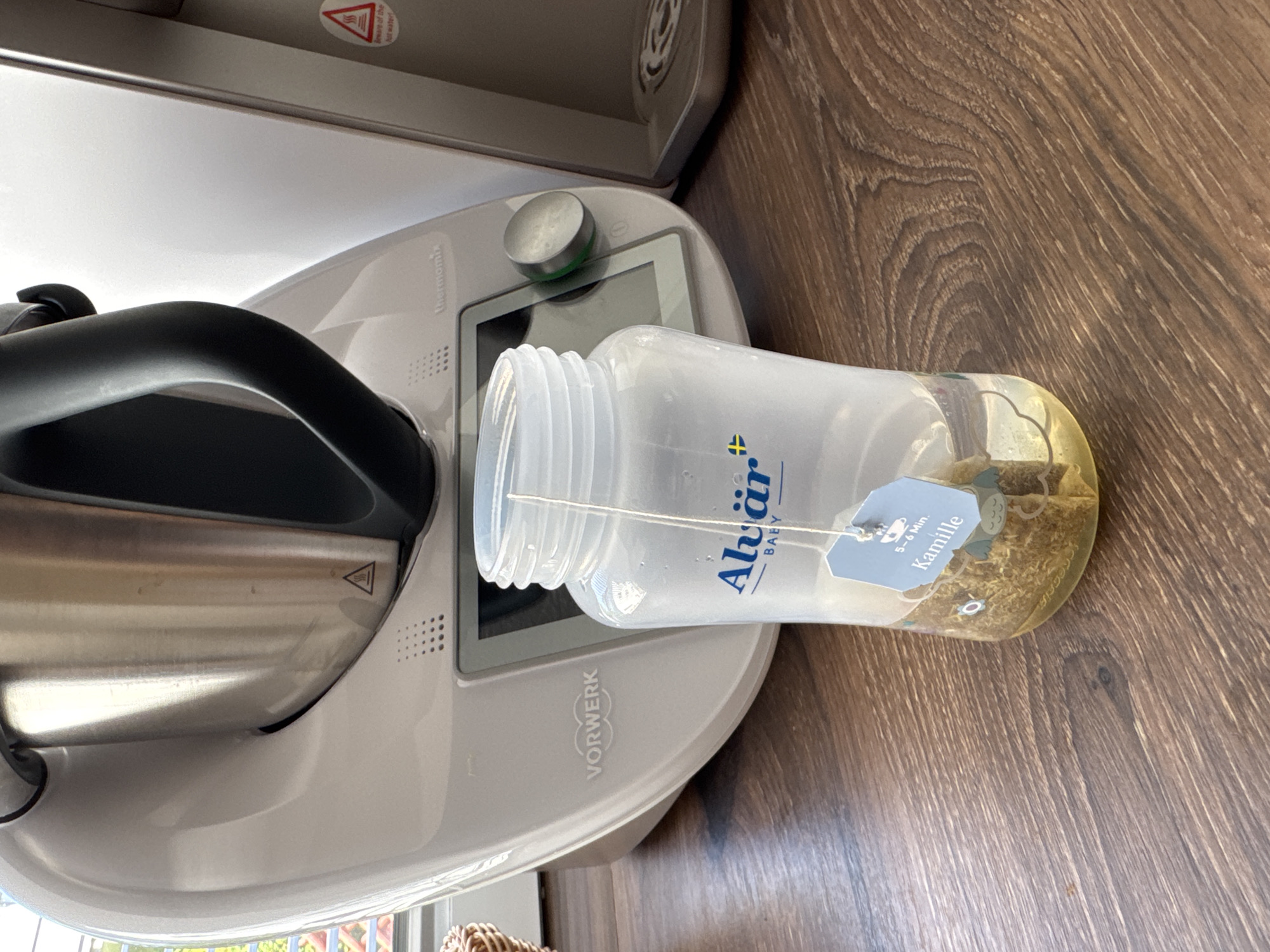 This media file is a close-up, angled shot of a white Thermomix kitchen appliance with a clear plastic bottle resting on it. The bottle contains water and dried chamomile tea leaves, with a tea bag tag visible that reads "Alvjår BABY" and "5-6 Min. Kamille". The Thermomix is on a wooden surface.
The appliance has a metallic bowl and a black handle. The bottle is positioned horizontally across the top of the Thermomix, with its opening resting inside the appliance's opening. The lighting suggests it could be daytime, but the specific time of day is not evident. There are no people visible in the image.
The scene appears to be in a domestic setting, possibly a kitchen, given the presence of the Thermomix and wooden surface. The focus is on the interaction between the Thermomix and the bottle of tea, suggesting the preparation of tea using the appliance. The overall mood is neutral and observational, focusing on the objects and their arrangement. There are warning symbols visible on the metallic part of the Thermomix, one depicting a flame and the other a triangle with an exclamation mark. The brand "Vorwerk" is also visible on the appliance.