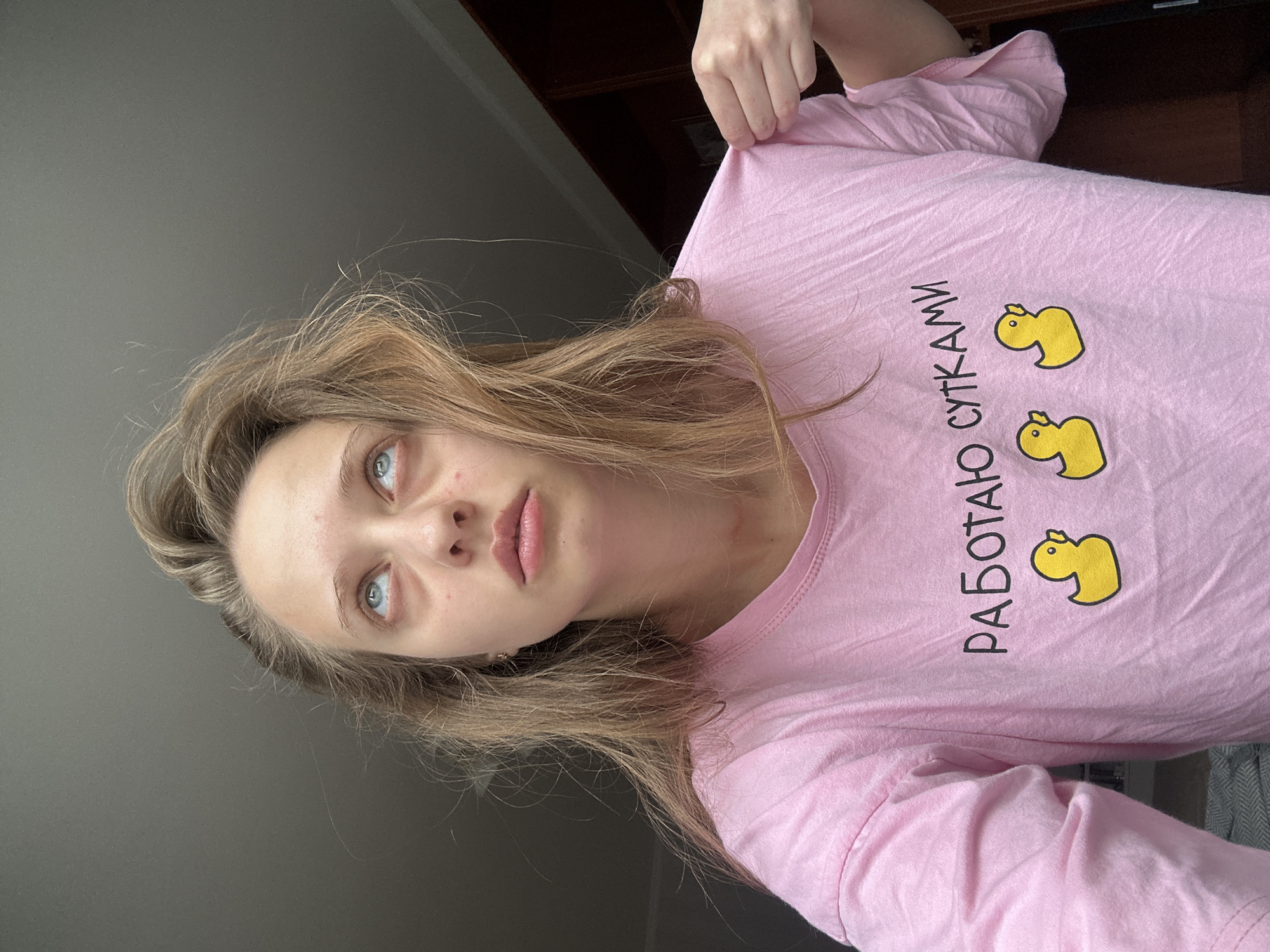 A young woman with blonde hair and blue eyes is looking up with a tired expression. She is wearing a pink t-shirt with a graphic of three yellow rubber ducks and text in Russian that reads "РАБОТАЮ СУТКАМИ" which translates to "WORKING FOR DAYS". Her hand is pulling on the edge of the t-shirt. The background appears to be an indoor setting with a window on the left and a wooden closet on the right. The lighting suggests it might be daytime. There is no discernible location in the city of Khimki, Russia.