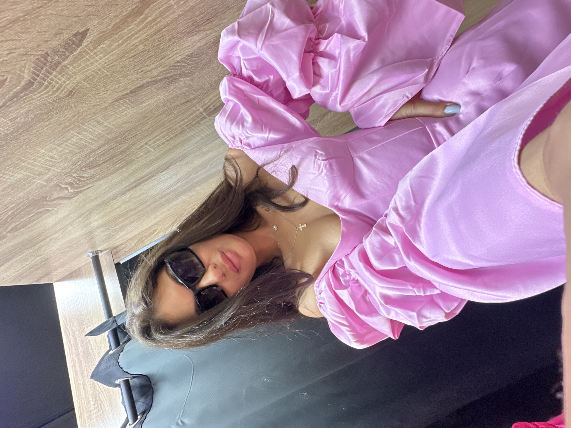 A person with brown hair and sunglasses is wearing a pink satin dress with puffed sleeves. They are lying down on a black surface, possibly a couch or bed, with their head near a light-colored wooden surface, possibly a table or floor. A chrome stand with a black bag is visible in the lower left corner of the image. The location is not determinable from the image alone, and there is no information to suggest it is Moscow, Russia.