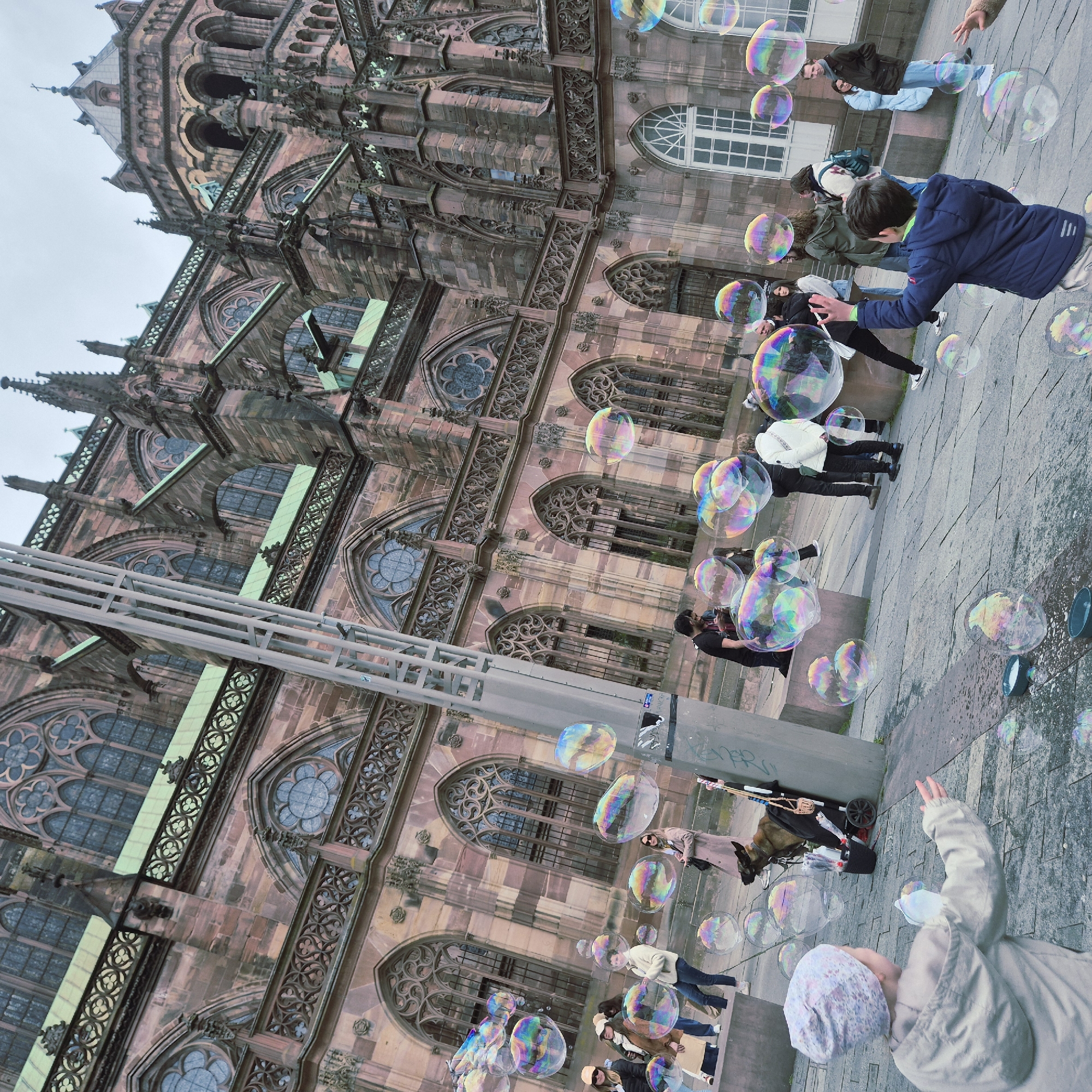 The image captures a scene outside the Strasbourg Cathedral in France. Several people are present on the paved area in front of the cathedral, with many individuals engaged in blowing and playing with large soap bubbles. The architecture of the Strasbourg Cathedral is visible in the background, showcasing its intricate stonework, arched windows, and spires. Some individuals are watching or attempting to interact with the bubbles. The overall environment appears to be a public space where people are leisurely enjoying the spectacle of soap bubbles near a historical landmark.