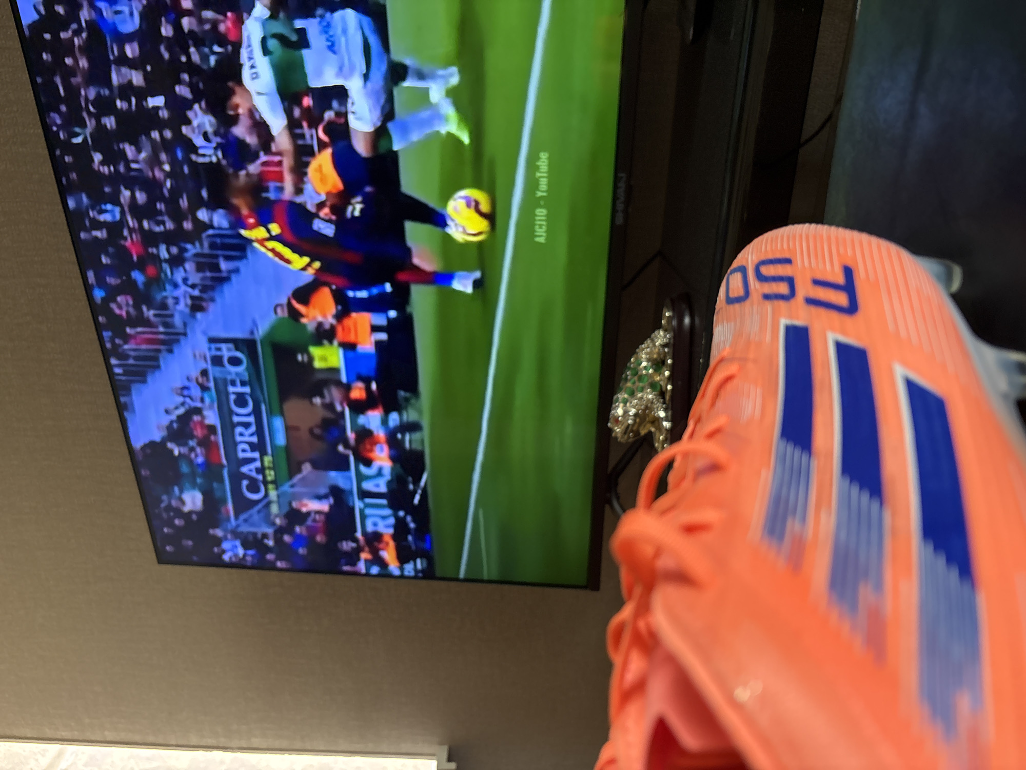 A television displays a soccer game. In the foreground is an orange Adidas F50 soccer cleat. The location is Yerevan, Armenia.