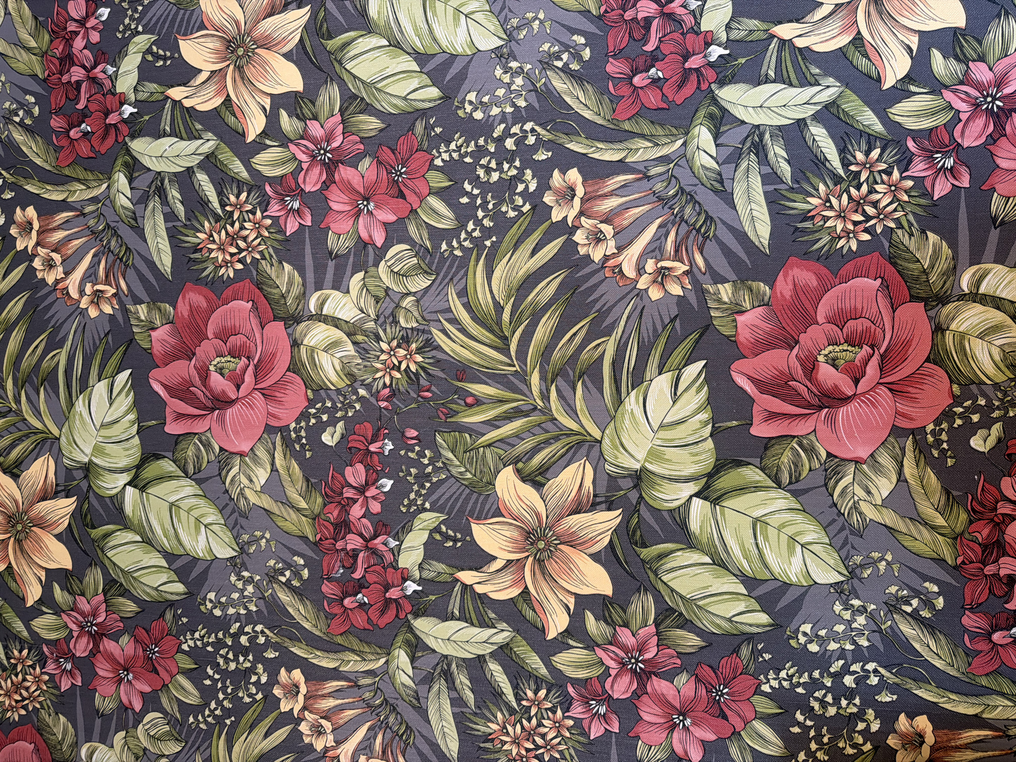 This is a close-up, top-down, full-frame, studio photograph of a dark gray fabric with a vibrant floral pattern. The pattern features a dense arrangement of stylized tropical flowers and large, elongated leaves in shades of pink, red, orange, yellow, and green. The flowers vary in size and shape, with some appearing like large, open roses and others like clusters of smaller blooms. The leaves are depicted with detailed venation and are shown in various orientations, overlapping each other and the flowers. The overall impression is one of a rich, tropical botanical print. The image is evenly lit, with no harsh shadows, suggesting controlled studio lighting.