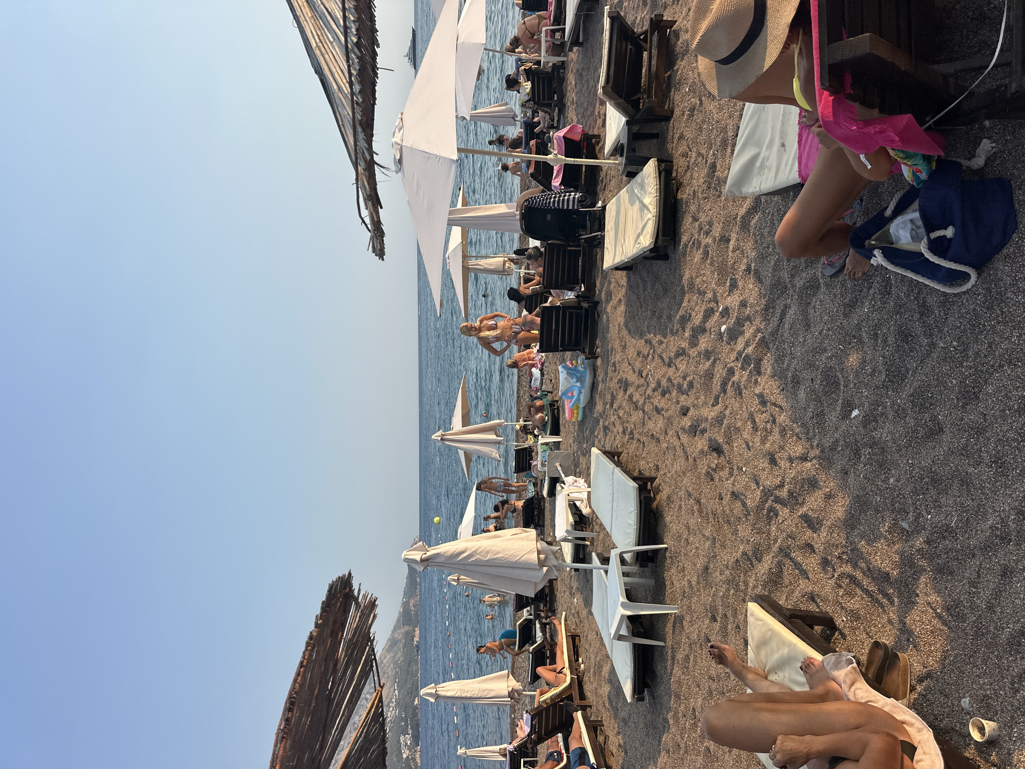This media file depicts a beach scene in Boreti, Montenegro, likely during the daytime, given the bright sunlight casting shadows. The setting is a pebble beach with numerous lounge chairs and umbrellas arranged for sunbathers. The sea is visible in the background, with calm water and a few people swimming or wading. Mountains can be seen in the distance, characteristic of the Montenegrin coastline.
Several individuals are present, mostly reclining on lounge chairs or walking along the shore. Some are wearing swimwear, suggesting a recreational activity like sunbathing or swimming. One person is standing and appears to be posing. The overall atmosphere is relaxed and leisurely, typical of a summer vacation day at the beach.
Notable details include the variety of beach furniture, such as white and wooden lounge chairs, and large white umbrellas providing shade. A dark blue beach bag and a straw hat are visible near one of the chairs. The sand or pebbles on the beach are dark in color. The lighting suggests it might be late afternoon or early evening, as the sun's angle creates long shadows. There is no visible text in the image.