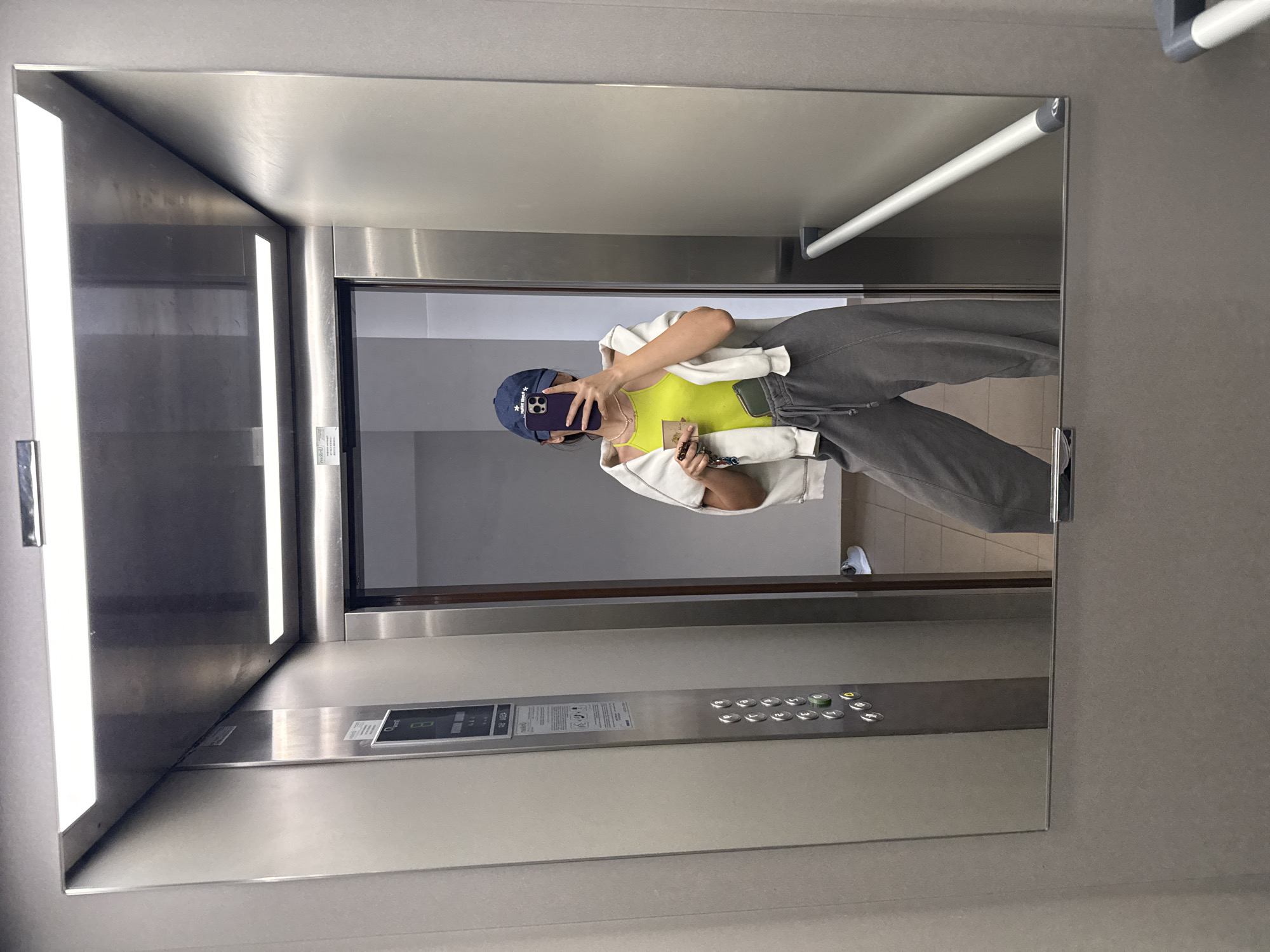 A person is captured in a mirror selfie inside what appears to be a modern elevator. The image, which is rotated 90 degrees clockwise from its natural orientation, shows the individual standing casually, holding a smartphone to obscure their face while taking the photo.
The person is dressed in a dark blue baseball cap, a vibrant neon yellow or lime green tank top, a white or cream-colored open hoodie or jacket, and grey sweatpants. Partially visible white sneakers complete the casual outfit. In their non-phone hand, they hold a set of keys with a small, light-colored charm and a slender wallet or cardholder.
The elevator's interior features sleek, brushed stainless steel panels and bright vertical fluorescent light strips that run along the edges of the mirror and the elevator entrance. A metallic handrail extends horizontally across the upper right portion of the mirror. On the elevator's right wall, a control panel is visible, equipped with numerous silver push buttons for floors numbered 0 through 14, as well as a 'P' button. Buttons for opening and closing doors, along with an alarm bell symbol, are also present. Some floor buttons, particularly '0', are illuminated green. Below the main button interface, several information labels and stickers are present, though most text is illegible, except for partially visible indications of "MAX" and "KG" likely detailing the elevator's capacity.
The setting suggests an indoor environment within a contemporary building in Plovdiv, Bulgaria. The lighting is artificial and bright. No specific time of day or weather is discernible from the indoor scene. The mood conveyed is casual, typical of a quick self-portrait in a public space.