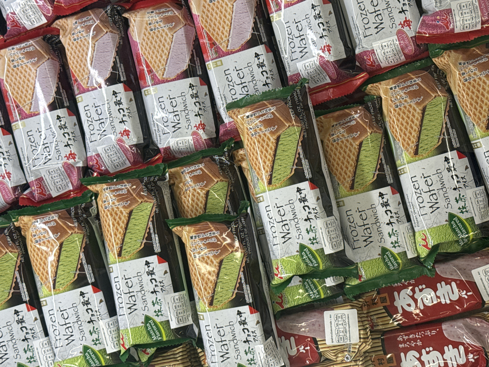 The image shows a close-up, overhead view of an assortment of frozen wafer sandwiches, primarily in green (matcha flavored) and pink (strawberry flavored) varieties, displayed in a retail setting, likely a freezer aisle in a supermarket in Bangkok, Thailand. The packaging features Japanese text and branding such as "Frozen Wafer Sandwich" and "抹茶チョコアイス" (Matcha Chocolate Ice). Some packages also display nutritional information in Thai and English. The lighting is bright, consistent with retail store lighting, and the overall scene is clean and organized, indicating a well-maintained display of frozen desserts.