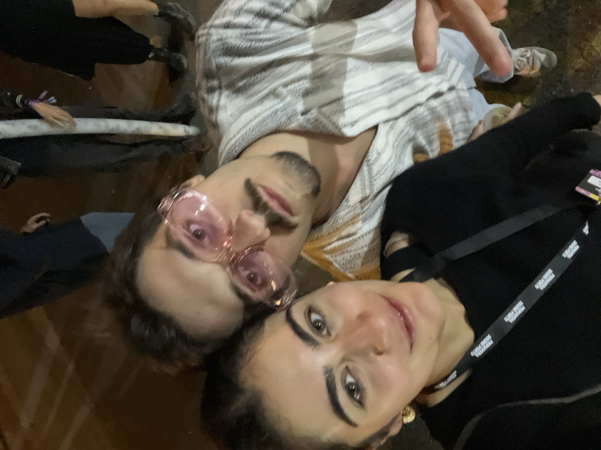 Two individuals are captured in a close-up selfie-style shot. The person in the foreground, on the left, is a woman with dark hair, wearing a black top and a golden hoop earring. A black lanyard with white text is visible around her neck. The second individual, to the right and slightly behind, is a man with a mustache and goatee, wearing pink-tinted glasses and a striped shirt. His right hand is raised, making a peace sign. The background appears to be a dimly lit outdoor setting with other people partially visible and a wooden surface on the ground. The context of the image, with the lanyard mentioning "PRIMAVERA SOUND PORTO," suggests the location is likely near the festival grounds in Matosinhos, Portugal, during the event.