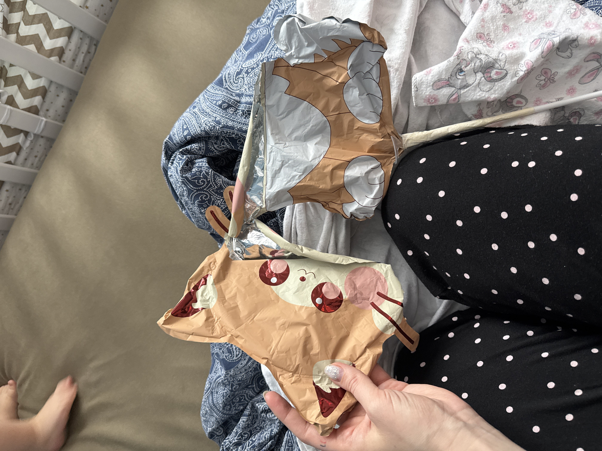 The image presents an overhead view of an indoor domestic scene, likely a bedroom or nursery, set in Moscow, Russia. The central focus is an adult's hand holding two deflated foil balloons. The lower balloon clearly depicts a cartoon animal face, possibly a deer or rabbit, with large red eyes, pink cheeks, and brown facial features, appearing to hold a red ice cream cone. The adult's hand, adorned with glittery nail polish, cradles this balloon.
The person is wearing black pajamas with white polka dots, and their upper body is covered by a blue fabric with a white paisley pattern. Adjacent to the person are light-colored fabrics, including a white one decorated with pink and grey cartoon bunnies (reminiscent of Thumper) and butterflies, suggesting a child's presence. A small child's foot is visible in the lower-left corner, resting on a beige mattress or sheet. To the far left, a white crib or bassinet with brown and white zigzag patterned bedding is partially seen. A white plastic straw, likely used for inflation, is inserted into one of the deflated balloons. The scene conveys a relaxed, casual atmosphere, possibly following a celebration or during playtime. The lighting is soft and ambient, indicating daytime. No visible text is present in the image.