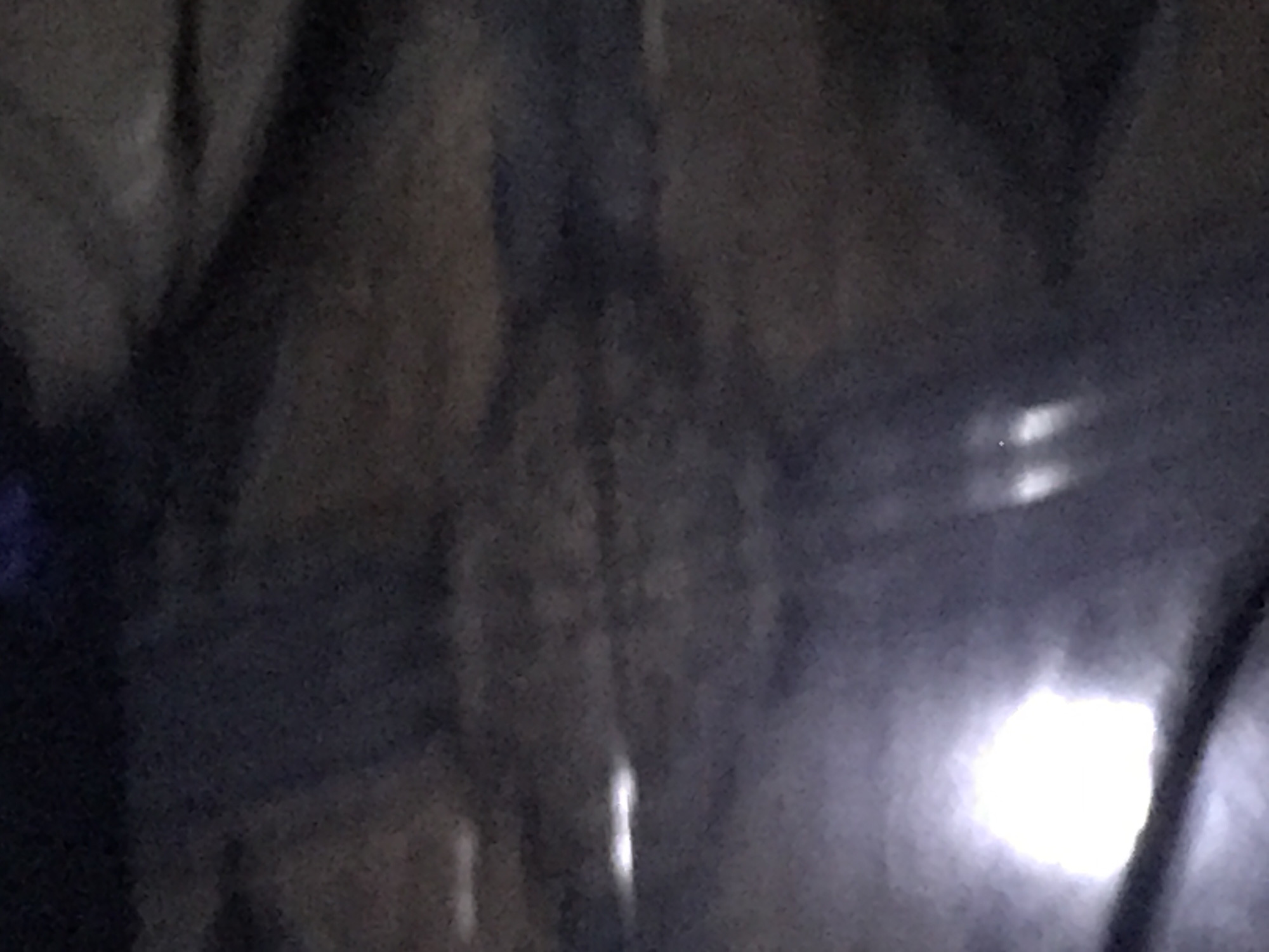 The image is an out-of-focus, dimly lit photograph that appears to have been taken at night or in low-light conditions. The scene is dark and hazy, with indistinct shapes and blurry light sources. In the lower left quadrant, there is a large, bright, white, amorphous light source, possibly a streetlamp or headlights. To the right of this, there are several smaller, elongated light streaks. The background consists of dark, muted tones with suggestions of horizontal lines or textures, possibly indicating a road or a textured surface. The overall impression is one of obscurity and a lack of clear detail.