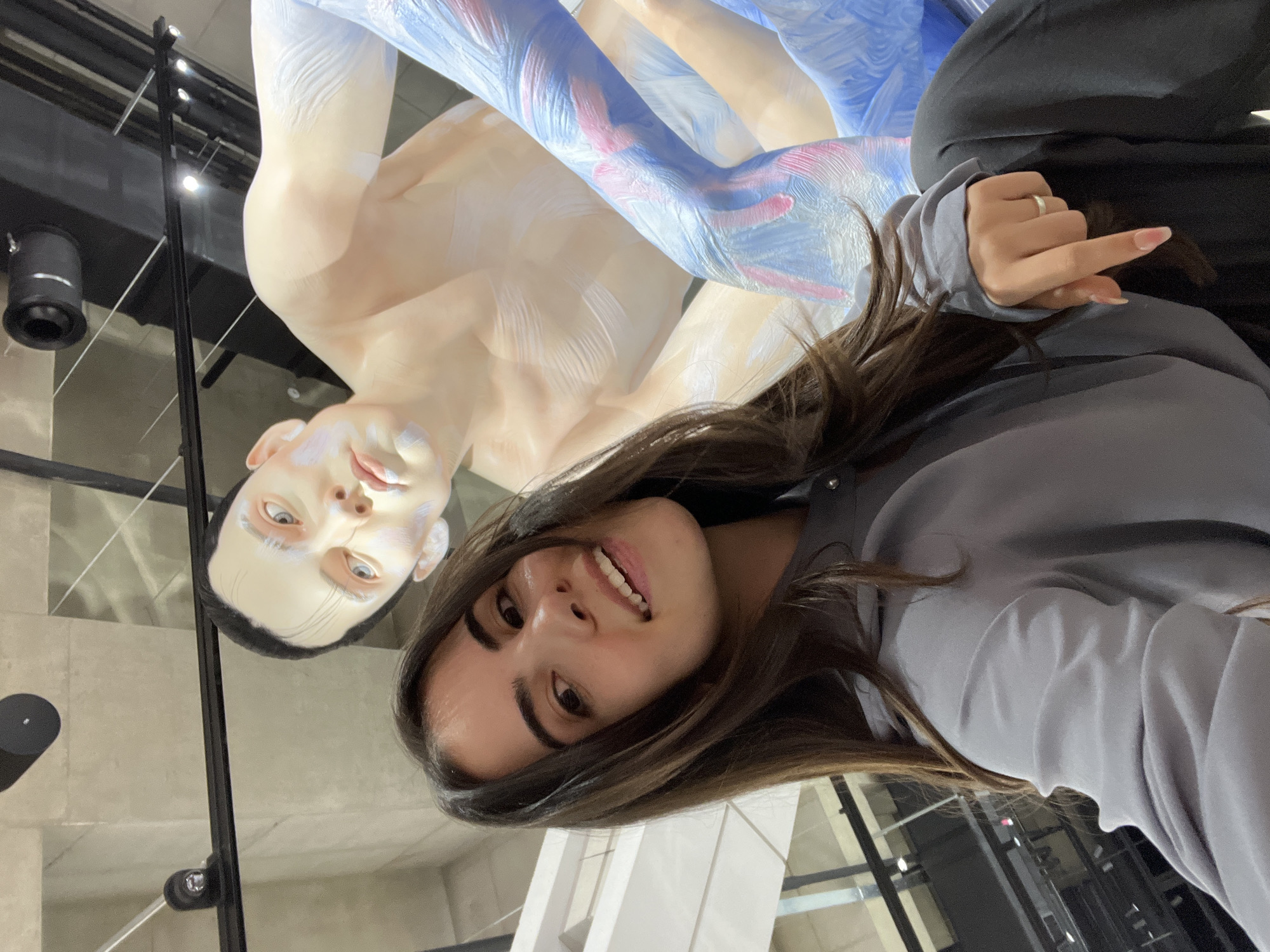 A young woman is captured in a selfie, smiling broadly at the camera in what appears to be an art gallery or museum in Seoul, South Korea. She has long, dark hair and is wearing a grey long-sleeved top. Her left hand, adorned with a ring on her index finger and neatly manicured fingernails, is visible in the bottom right of the frame.
Behind and above her is a large, artistic sculpture of a human figure. The sculpture is predominantly pale or light-toned, possibly depicting a male form, and features abstract blue and pink paint strokes across its limbs and torso, giving it a stylized appearance. The sculpture's face has pale eyes and is looking upward and slightly away from the viewer.
The background indicates a modern indoor setting with high ceilings, artificial lighting, and contemporary architectural elements such as concrete surfaces and black support structures. The woman's expression is joyful and engaged, suggesting she is enjoying her visit to the exhibition. No text is visible in the image.