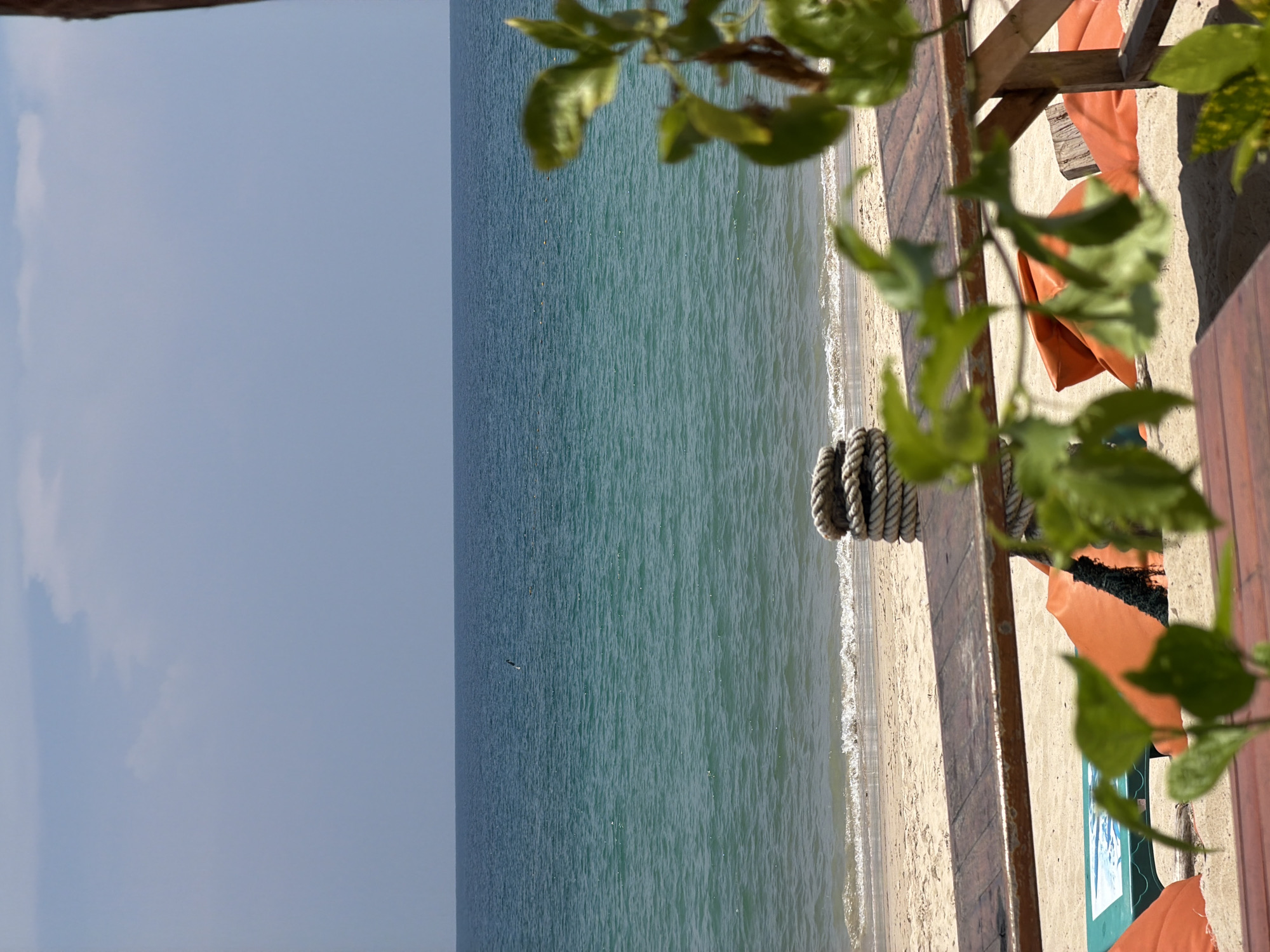 The image presents a serene, sunny view of a tropical beach and ocean, likely from a raised wooden platform or deck, possibly in Choeng Thale, Thailand.
In the foreground, lush green foliage with somewhat blurry leaves and branches partially frames the upper right and bottom right of the scene, suggesting the presence of nearby trees or plants. A sturdy wooden structure, possibly a railing or part of a deck, runs along the right side of the frame. On the sandy area adjacent to this structure, a thick, braided rope is coiled, resting on the sand. Further into the scene, several orange, beanbag-like lounge chairs are visible on the light-colored sand, arranged for relaxation. A small green object, possibly a cooler or low table, with a blue and white design is partially visible near the chairs.
The midground is dominated by a strip of sandy beach where gentle, white-crested waves lap at the shore, creating a foamy edge along the waterline. Beyond the beach, the vast expanse of the ocean stretches out. The water is a beautiful clear turquoise-green, appearing calm with subtle ripples across its surface. The distant horizon line is visible, separating the sea from the sky.
The background features a clear, light blue sky with only a few faint, wispy white clouds. The overall impression is one of a peaceful, bright day, suitable for leisure and enjoying the coastal environment. There are no people visible, and no discernible text appears in the image.