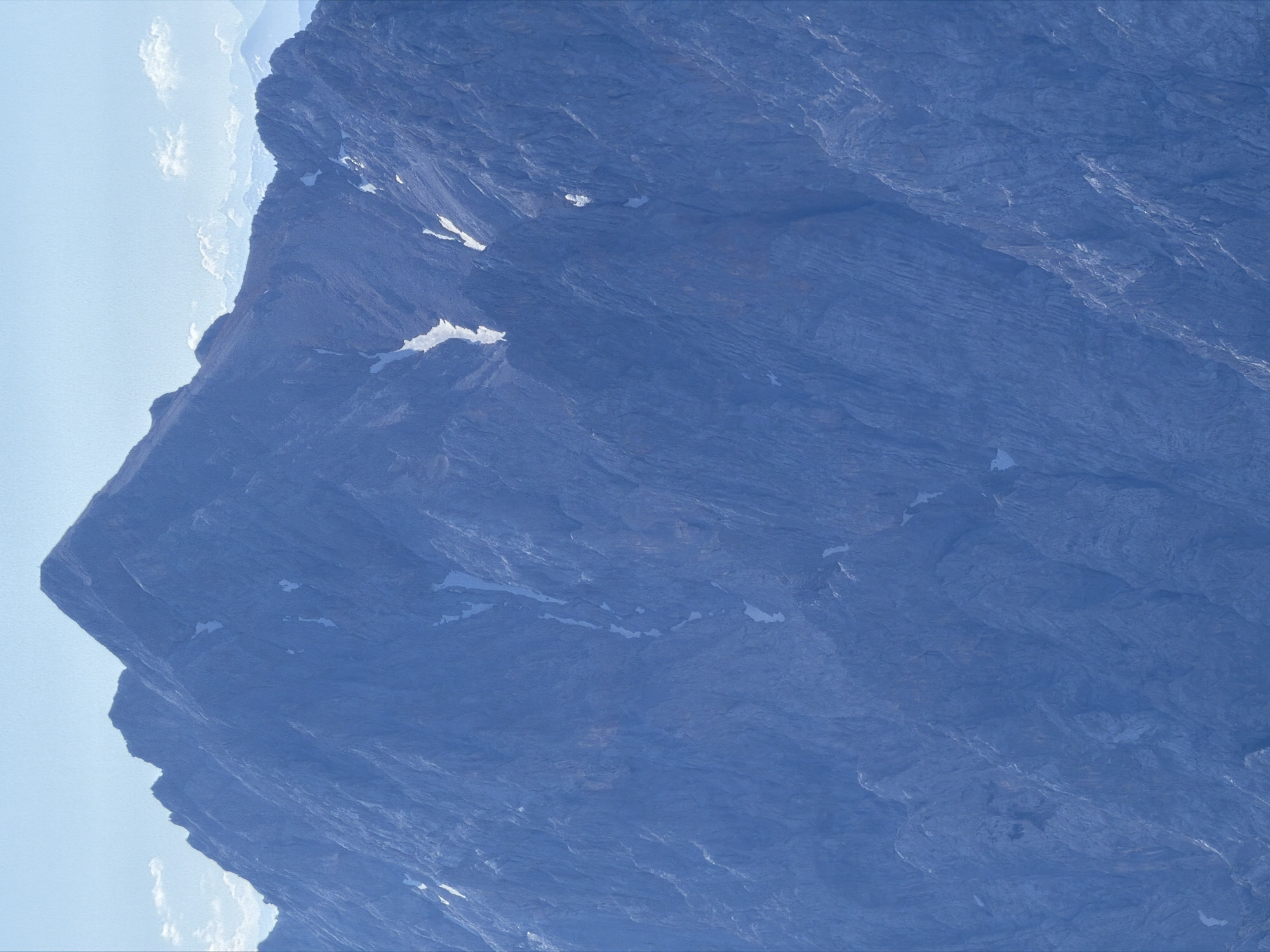 The image shows a close-up view of the Zugspitze mountain, the highest peak in Germany, located near Garmisch-Partenkirchen. The mountain is predominantly blue and gray, with rugged, rocky terrain. Patches of snow are visible in crevices and on the upper slopes, contrasting with the dark rock. The sky above is a pale blue with a few wispy clouds. The overall impression is one of a vast, imposing natural landscape.