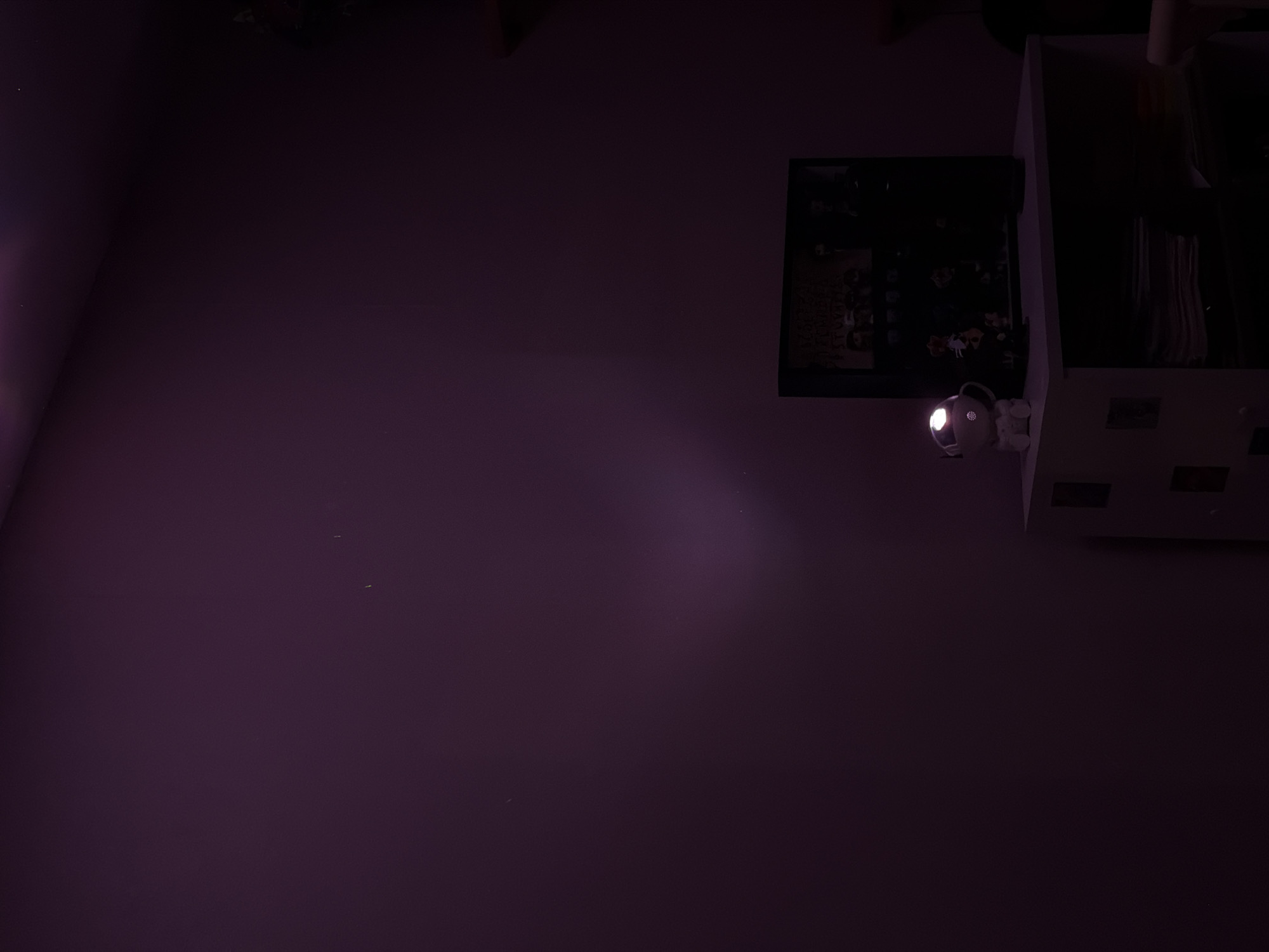 The scene presents a dimly lit interior space. Dominated by a wall surface in a purple hue, the frame captures an overhead perspective. A framed image, seemingly a poster or print with people depicted, hangs near the upper right. Adjacent to it is a white, box-like structure featuring vents. A small spotlight is mounted near the bottom center of the image, casting a beam of light on the wall. The environment appears residential, with limited visible details. The location in the city of Perm, Russia, is not discernible from the image.