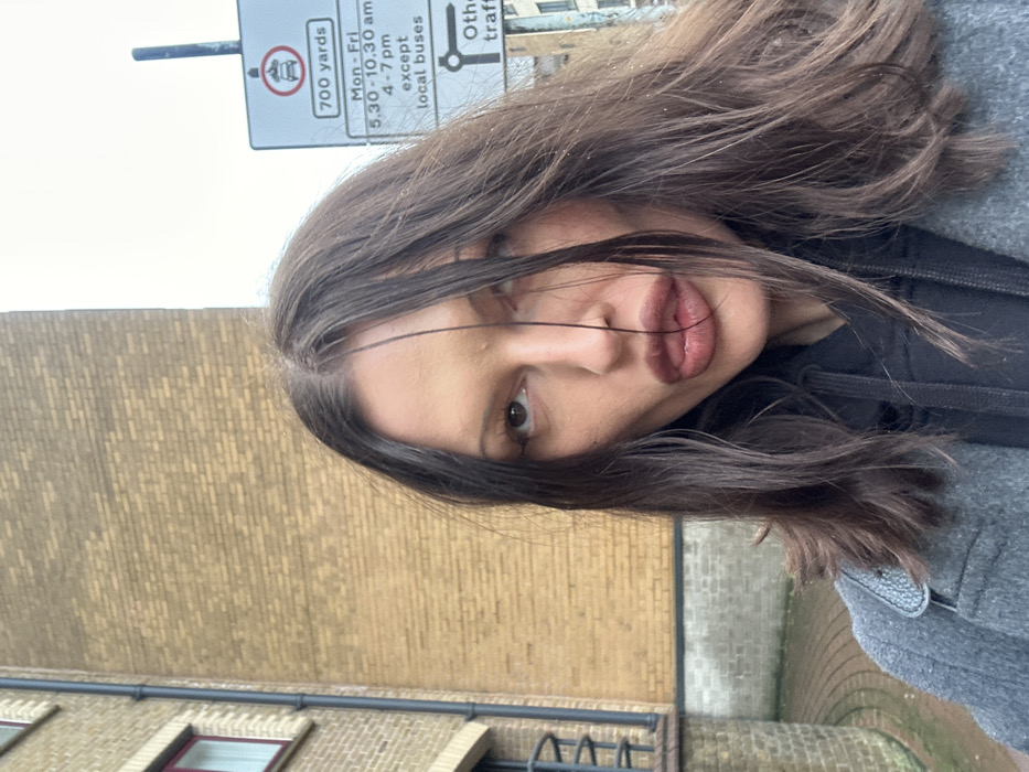 An adult female is captured in a close-up outdoor portrait, her head and upper torso filling the majority of the frame. She possesses dark, shoulder-length hair, centrally parted, with strands falling across her face. Her full lips are slightly pursed, and her eyes gaze upwards and to the right. She is dressed in a grey coat layered over a black hooded garment.
The background reveals urban infrastructure. To the left, a building constructed of light brown brickwork is visible, featuring a dark vertical drainpipe and a window with a dark red frame. The ground beneath this building shows curved grey brick paving. To the right, a white rectangular road sign, mounted on a dark pole, is partially visible. The sign displays a red circular symbol prohibiting motor vehicles, accompanied by the text "700 yards". Below this, specific time restrictions are detailed: "Mon - Fri", "5.30 - 10.30 am", and "4 - 7 pm", with an exception noted as "except local buses". A partial arrow pointing right with "Other traffic" is also visible. The specific typography, symbols, and traffic restriction parameters on the sign are characteristic of the United Kingdom, placing the scene in the city of London. The ambient lighting appears overcast. No other individuals or active interactions are observed.