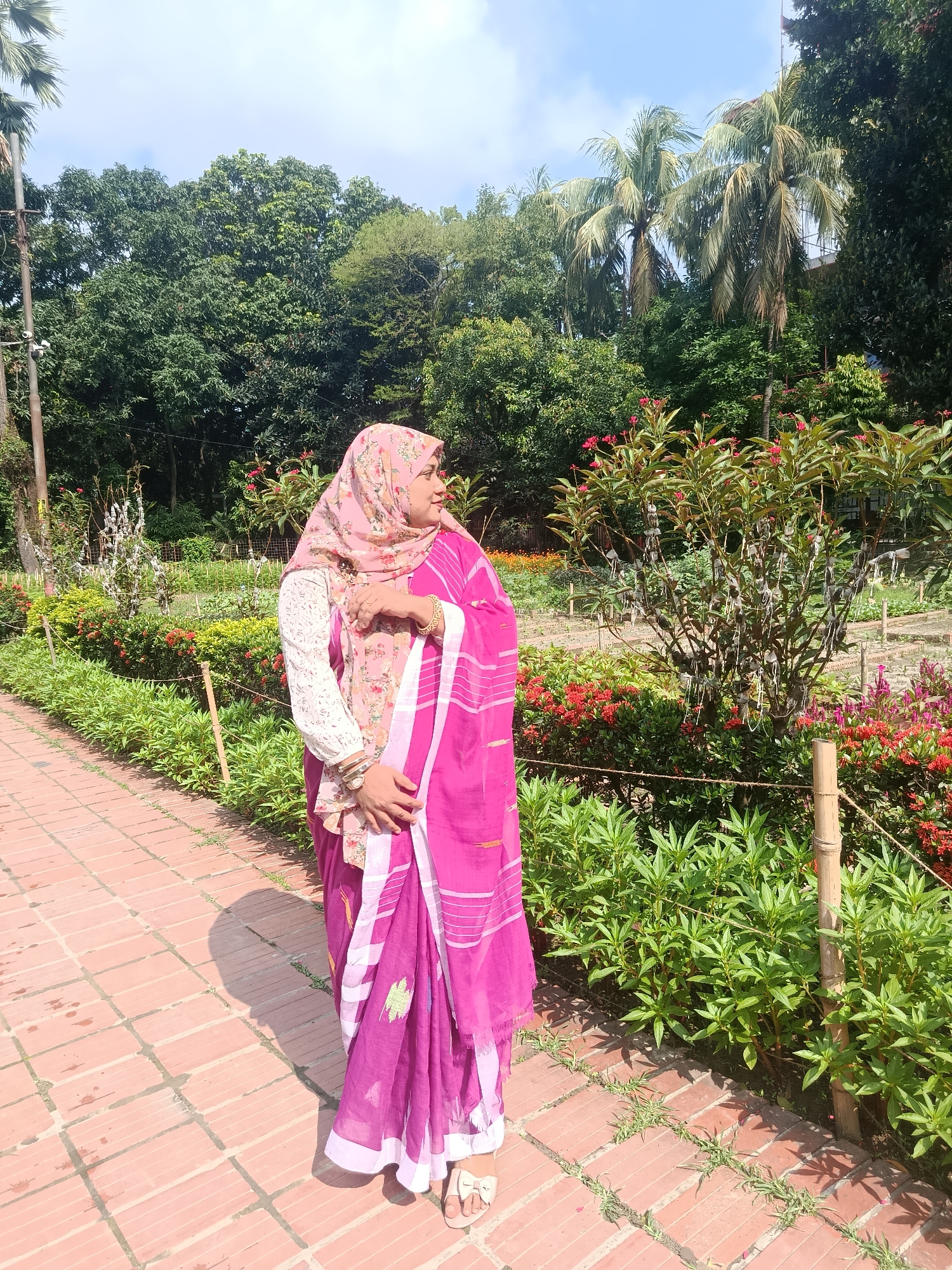 A woman wearing a pink sari and a floral hijab stands on a brick pathway in a garden. The garden has a variety of trees, including palm trees, and flowering plants with red blooms. The pathway is lined with low-lying greenery. The sky is partially visible in the background. The location is likely in Dhaka, Bangladesh, based on the clothing style and environment.